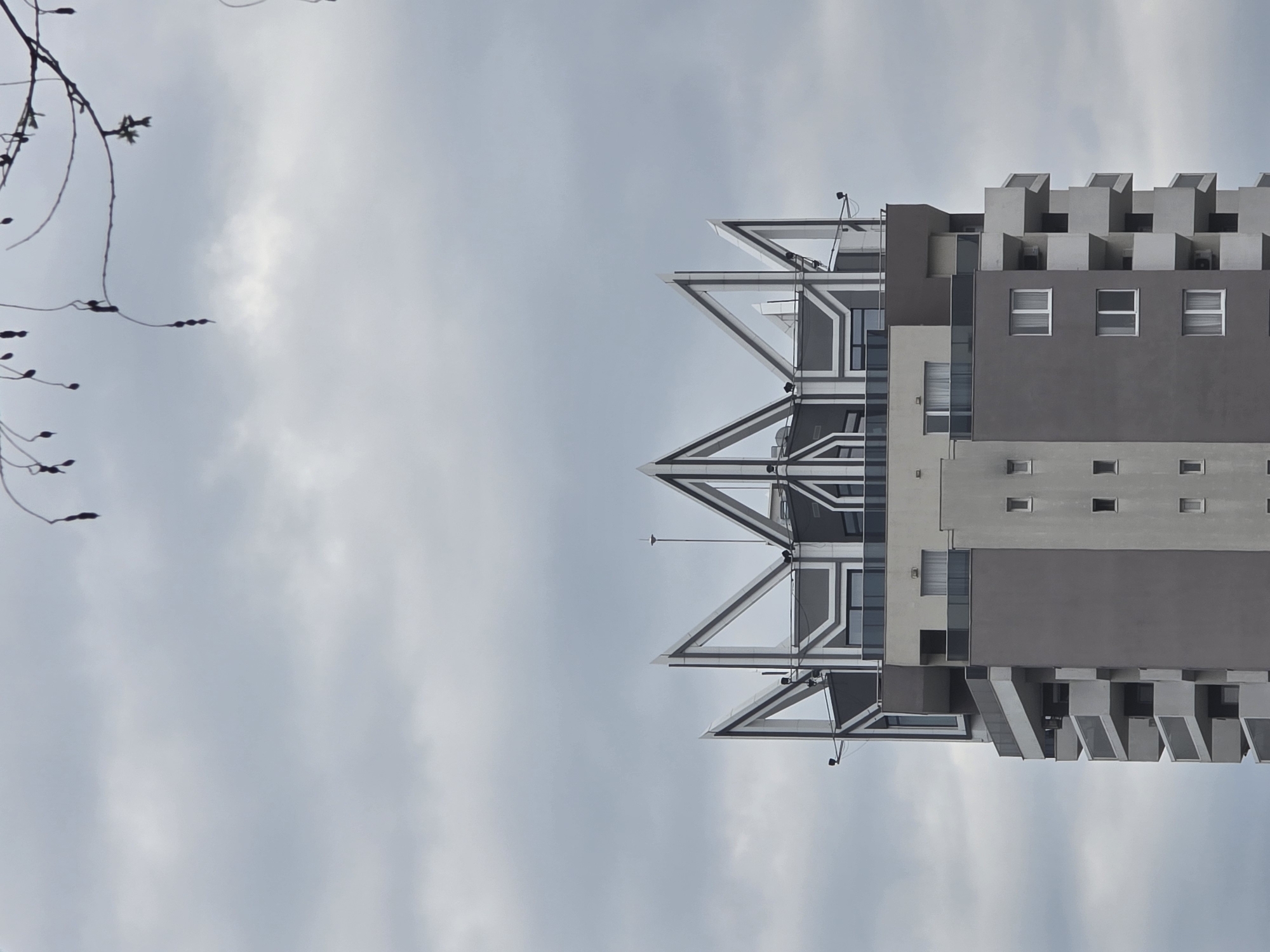 The image shows a tall building in an unknown city. The building has a unique design, with a series of triangular structures on its top. It also has several windows and balconies. The sky is cloudy, and there is a tree branch visible in the left corner of the image.