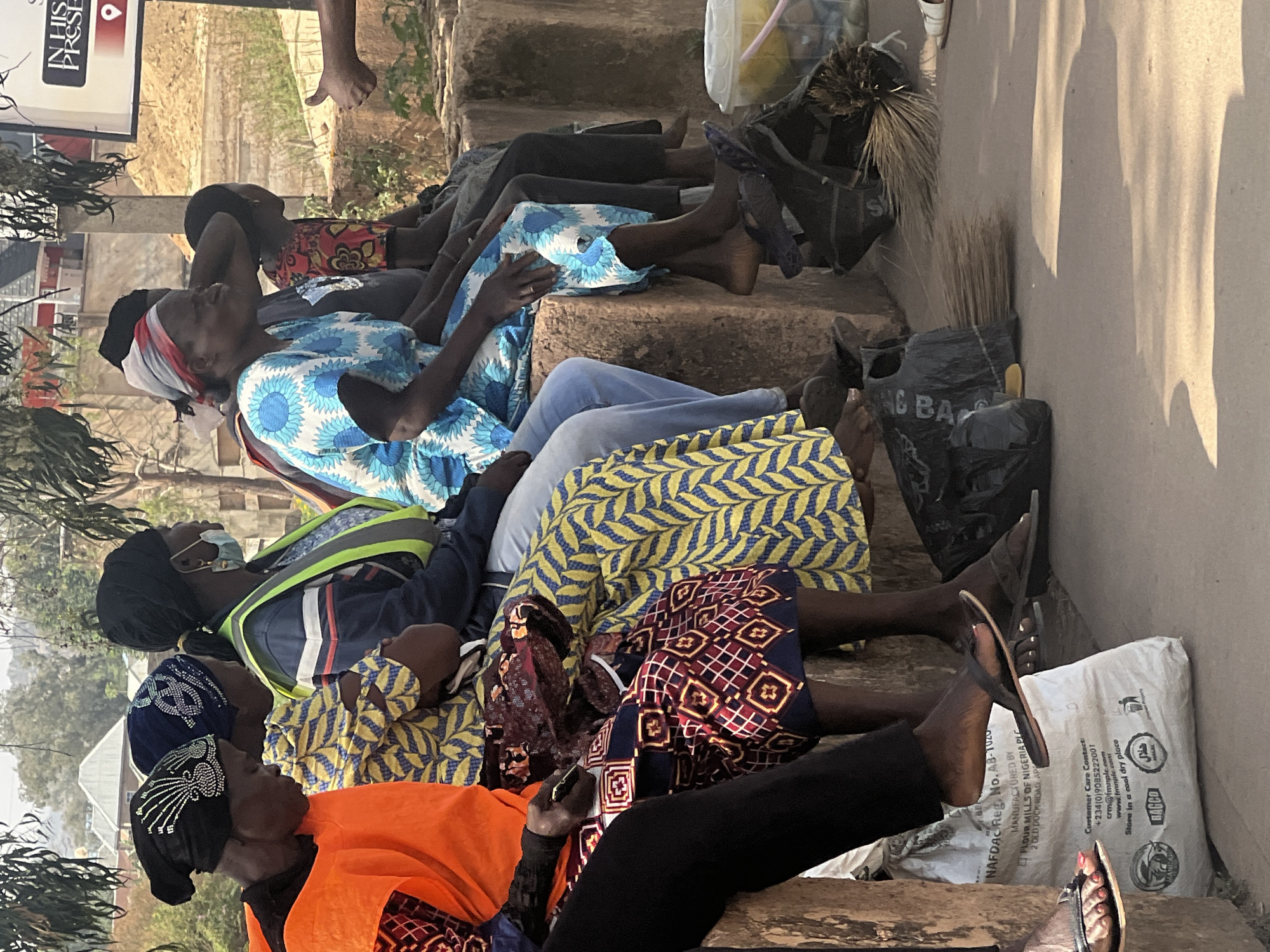 A vertically oriented image depicts a group of people, primarily women, seated on concrete steps or ledges in an outdoor setting in Jos, Nigeria. The scene is bathed in sunlight, casting distinct shadows on a light-colored wall to the right, indicating daytime.
The individuals are positioned in a line, appearing to be resting or waiting. Their clothing features a mix of traditional patterns and vibrant colors; notable attire includes a blue and white floral dress, yellow and black geometric prints, and a bright orange top. One person wears a dark blue jacket with a high-visibility yellow-green vest and a light blue face mask. Most are either barefoot or wearing simple sandals.
Various objects are present, including a prominent white bag on the right, which appears to be a flour sack. Visible text on this bag states "NAFDAC Reg. No. AB-1020", "MANUFACTURED BY FLOUR MILLS OF NIGERIA PLC", "Customer Care Contact: +234(0)9085222001", "crm@fmnplc.com", "www.fmnplc.com", "Store in a cool dry place", "BAGCO", and the Arabic "حلال" (Halal) symbol. Black plastic bags containing brooms made of natural fibers are also visible, one partially showing "G BAG". In the upper left, a white sign with a red and white accent displays "IN HIS PRESE" (likely "PRESENCE") and a location pin icon. The background shows sparse trees and a dusty, open area. The overall impression is one of calm and daily life in a local community.