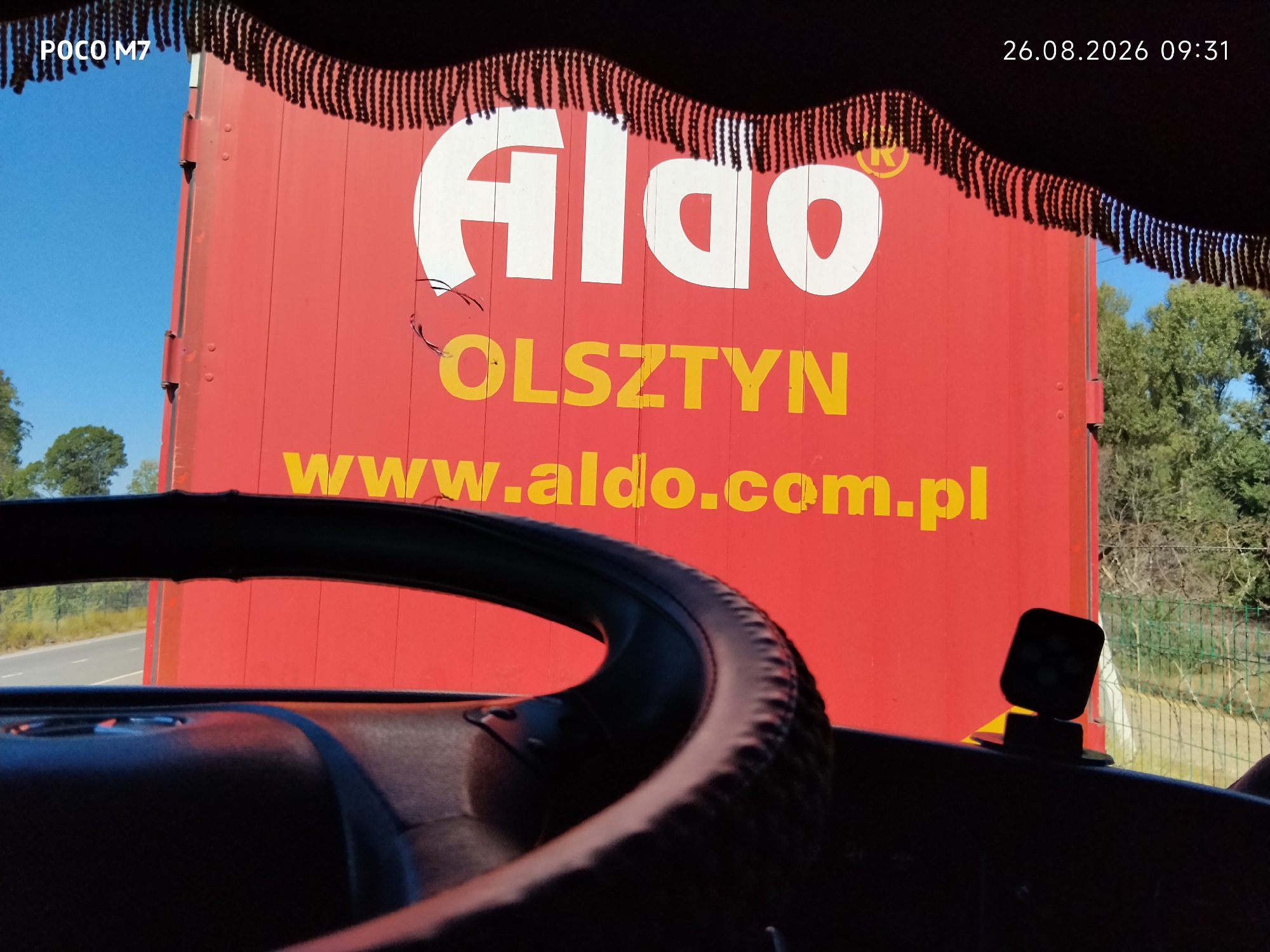 The image, taken on August 26, 2026, at 09:31, shows a view from inside a vehicle, looking out at the side of a large red truck trailer. The trailer has a white logo that appears to read "ALDO" with a registered trademark symbol. Below the logo, in yellow letters, is "OLSZTYN" and then "www.aldo.com.pl". The top of the trailer is adorned with a fringe of black decorative elements. Outside the trailer, the sky is blue, and there are green trees visible on the right side, along with a fence. The left side of the image shows a road and some vegetation. The interior of the vehicle includes a portion of the steering wheel and dashboard. The location in the city of Ауқатты, Kazakhstan, is not determinable from the visual information in the image.