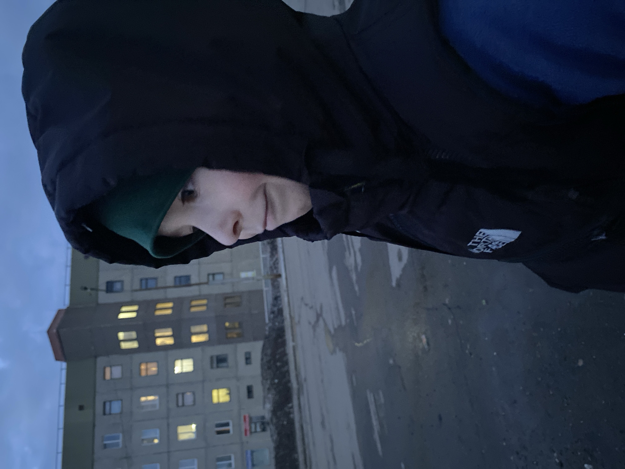 A person in a dark hooded jacket and a green beanie stands in front of a tall, gray apartment building in Марковский, Russia. The building has a distinctive red-brown section at its top and numerous windows, many of which are illuminated. The ground appears to be wet or covered in snow. The photo is taken at an angle. The person is partially facing away from the camera.