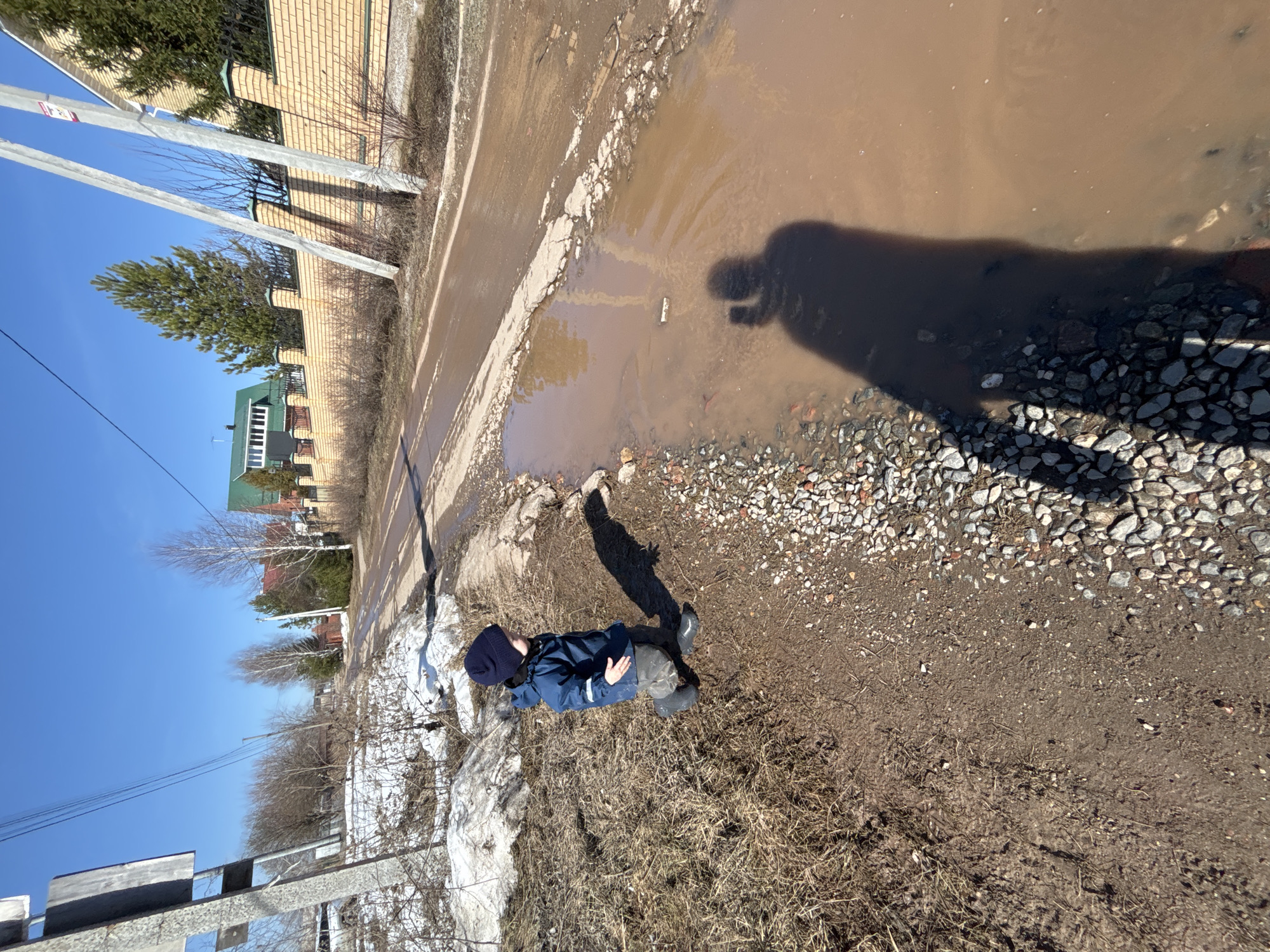 A vertical, medium shot photograph depicts a dirt and gravel roadside in daylight, with a child standing near a large puddle. The child, dressed in a dark blue jacket and hat, is standing sideways with their shadow cast on the puddle and adjacent gravel. The puddle is brown and murky. The roadside consists of a mix of dirt, gravel, and sparse vegetation. In the background, a damaged road edge is visible next to a brick wall. Beyond the wall, there is a building with a green roof. Trees and power lines are also visible. The location is Ферма, Russia based on the query. Remnants of snow are present on the lower left corner.