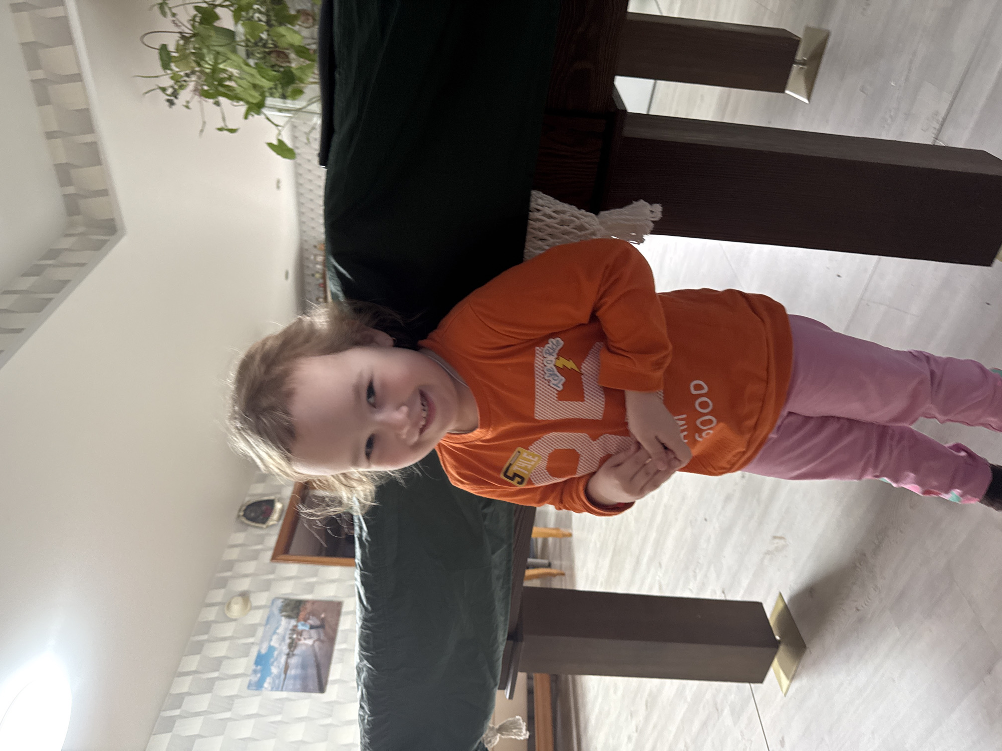 A young child in an orange long-sleeved shirt and pink pants stands next to a dark wood billiard table covered in green felt. The child is smiling and holding their hands together. The location is Zhigalki, Russia, inferred from other available data. Behind the billiard table and child is a white wall with decorative molding and framed pictures. The floor is a light-colored wood. A green potted plant is visible in the background, near a window.