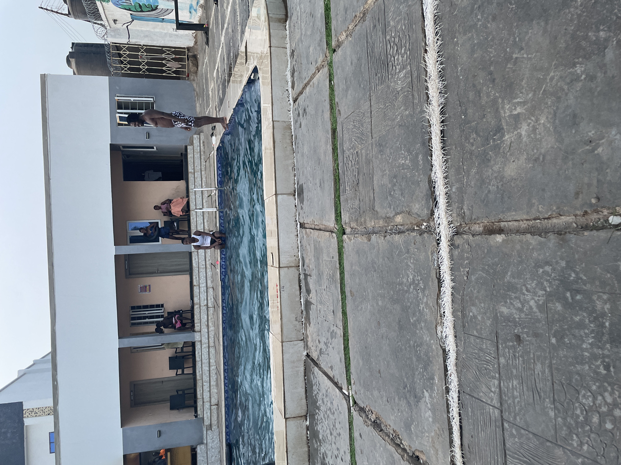 The image depicts an outdoor swimming pool area at a hotel or resort in Akwanga, Nigeria. The pool is surrounded by concrete paving stones with artificial turf in the joints. A two-story building runs alongside the pool, with multiple rooms visible, some with chairs and people. At the edge of the pool, a person wearing swimming trunks is preparing to jump in. Two other individuals are partially visible in or near the pool. The sky is light and clear. The building appears to have water tanks and security measures (barbed wire) on the roof.