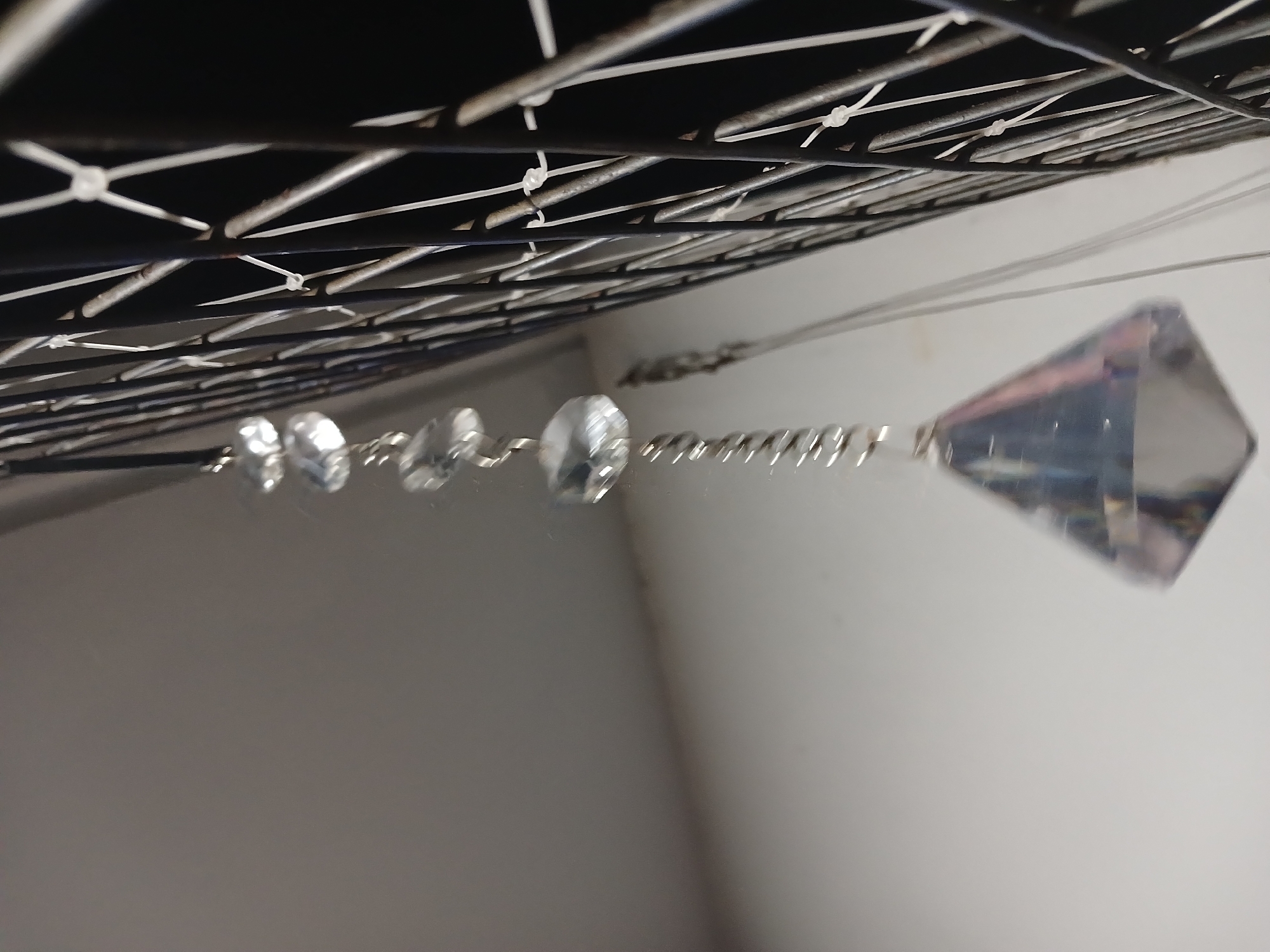 The image shows a crystal prism hanging from a wire grid with metal bars. The prism is elongated and has a pointed end. It is attached to the grid with a thin wire. The wire has small, clear beads attached to it before connecting to the prism. The background appears to be a white wall. The picture was not taken in Buenos Aires, Argentina.
