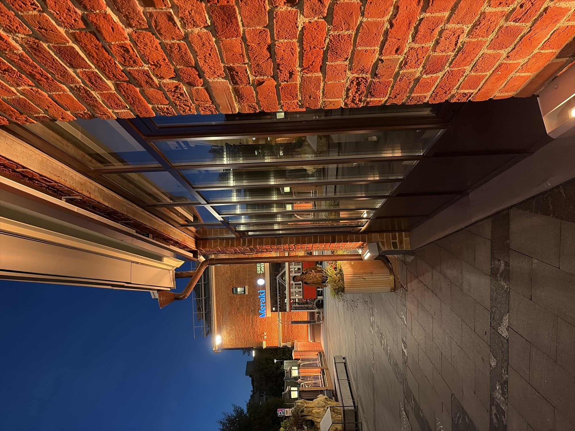 The image captures an outdoor scene at dusk in Moscow, Russia, specifically near the Meraki Green Tavern. A brick building with large windows is prominently featured on the right side of the frame, its brickwork illuminated by warm orange light. To the left, a paved walkway leads towards the building, with several street lamps and a large planter box visible. In the background, the Meraki Green Tavern signage is illuminated in blue. A person wearing a yellow jacket is walking away from the camera on the walkway. The sky is a deep blue, indicating late evening.