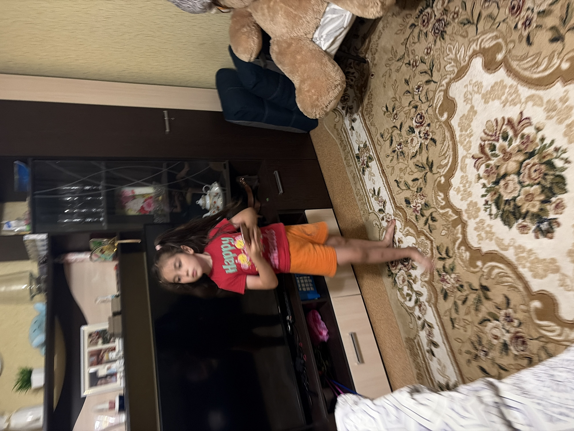 A young girl with dark hair in pigtails stands barefoot on a patterned rug in a dimly lit room. She is wearing a red t-shirt with the word "Happy" printed on it and orange shorts. She is holding a smartphone in her hands. To her left is a black television set on a dark wood entertainment unit. To her right, a large, light brown teddy bear sits on a chair. Behind the girl is a tall, dark wood cabinet with glass doors, and further to the right, a textured yellow wall. The room appears to be a living room.