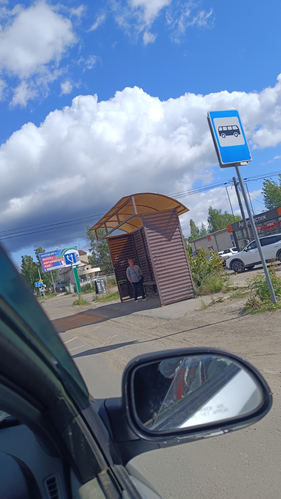 The image depicts a bus stop in Chaikovsky, Russia. A woman wearing a striped shirt and dark pants stands under the covered shelter of the bus stop. A blue and white bus stop sign is visible in the upper right corner. In the background, there are buildings, trees, and cars, including a white SUV. The scene is captured from the perspective of someone inside a vehicle, with a side mirror reflecting the interior of the car. The sky is partly cloudy with large white cumulus clouds. There is also a billboard in the background with Russian text.
