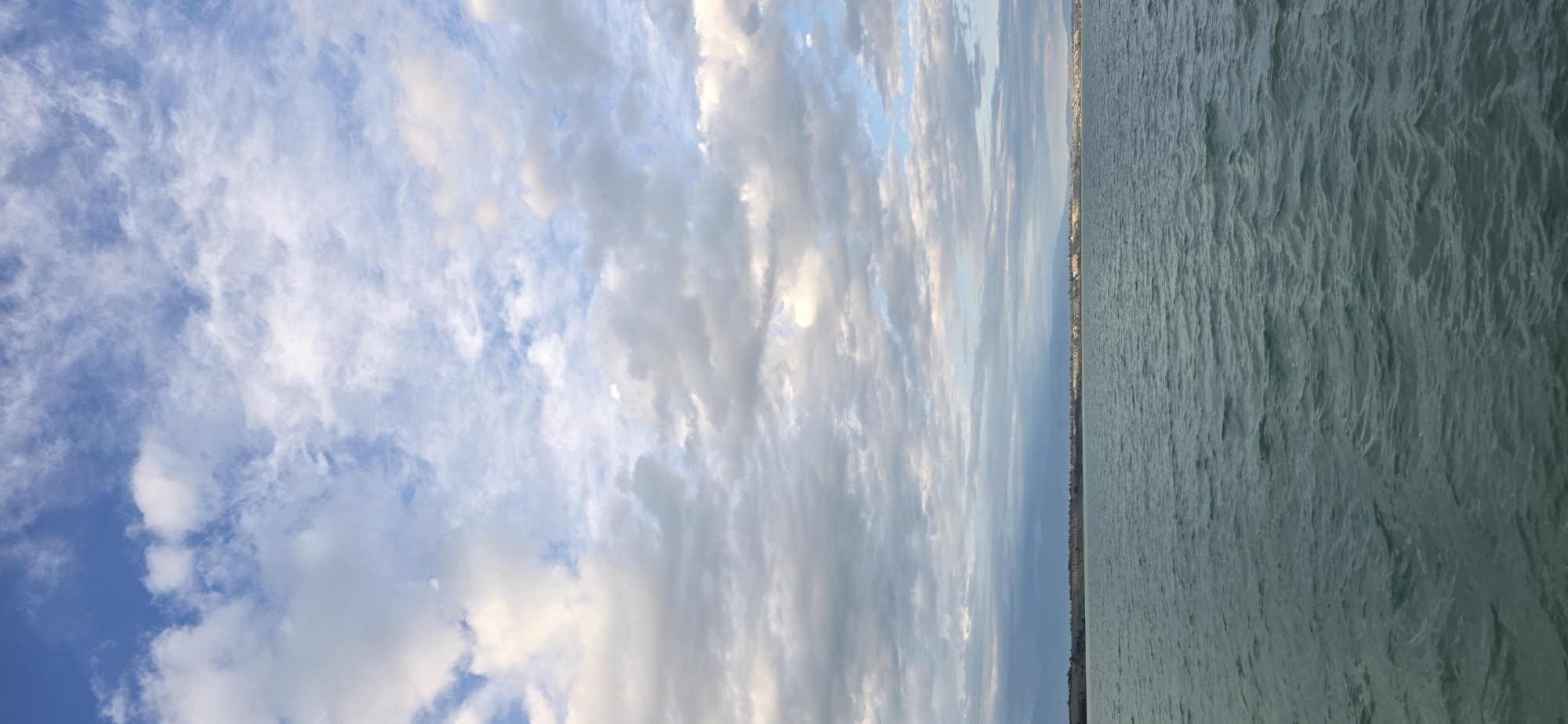 The image depicts a wide expanse of choppy, teal-colored water in the foreground, reflecting the sky above. The horizon line is visible, separating the water from a distant, flat shoreline. Beyond the shoreline, a range of muted, mountainous terrain stretches across the background, partly obscured by atmospheric haze. The sky is filled with a variety of clouds, ranging from scattered, wispy white clouds higher up to denser, grayer formations closer to the horizon. The lighting suggests it may be late afternoon or early evening, with a soft, diffused light illuminating the scene. The overall impression is one of a serene natural landscape, likely a lake or a large body of water. The city of Pristan'-Przheval'sk, Kyrgyzstan, is situated on the eastern shore of Lake Issyk-Kul, which aligns with this visual description.