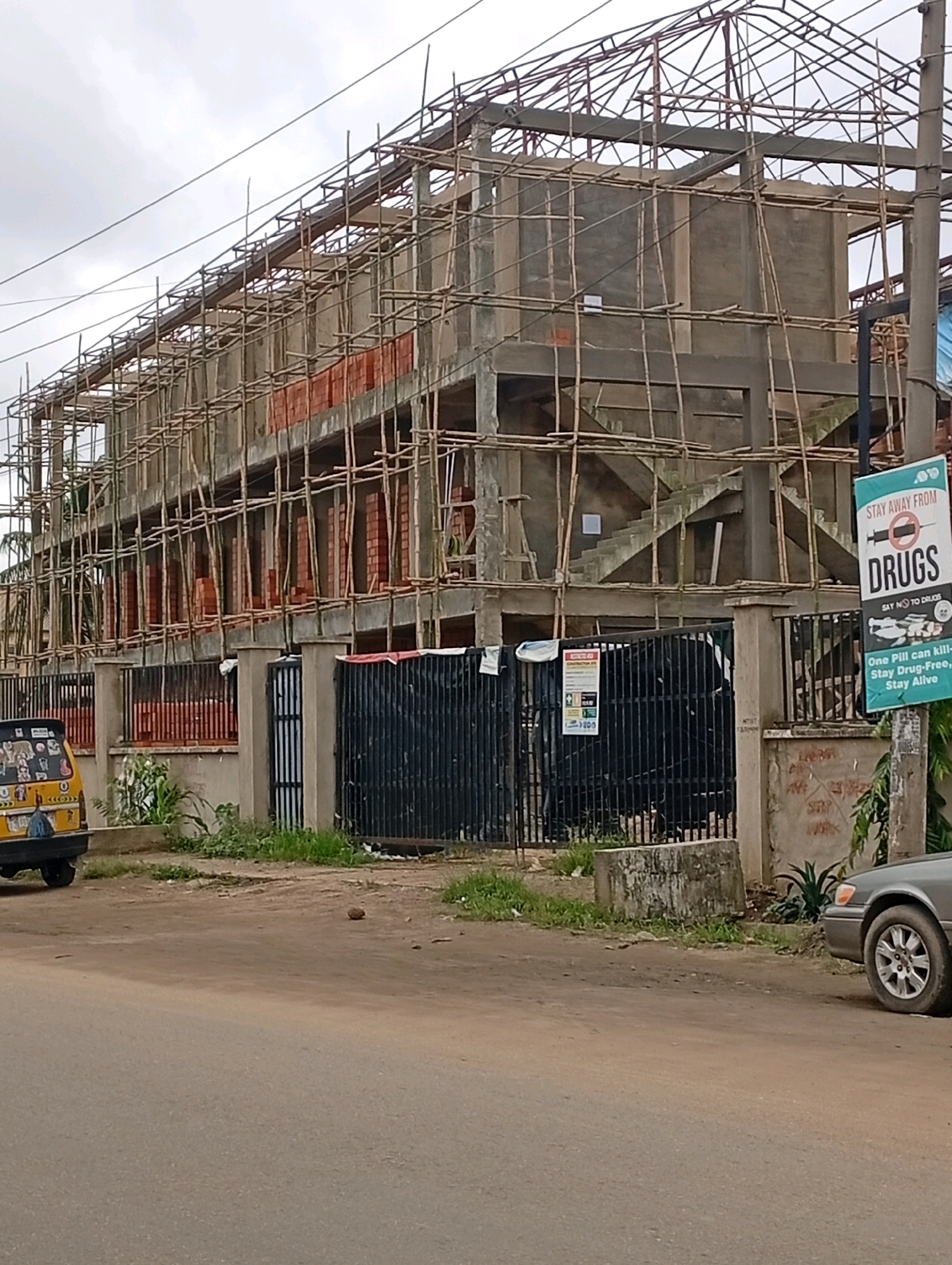 The image depicts a multi-story building under construction in Akowonjo, Nigeria. The building is partially constructed with concrete columns and floors, and a metal framework for the roof is visible. Bamboo scaffolding surrounds the structure, indicating ongoing construction work. Red bricks are stacked in sections along the exterior walls. In the foreground, a yellow motorized tricycle, often used as public transport in Nigeria, is parked on the dirt road. Black metal gates and a concrete wall separate the construction site from the road. A sign with the message "STAY AWAY FROM DRUGS" and promoting a drug-free lifestyle is posted on a pole near the entrance to the site. A gray sedan is parked further down the road on the right side of the image. The environment appears to be a roadside urban setting, with some greenery and debris scattered around. The sky is overcast.