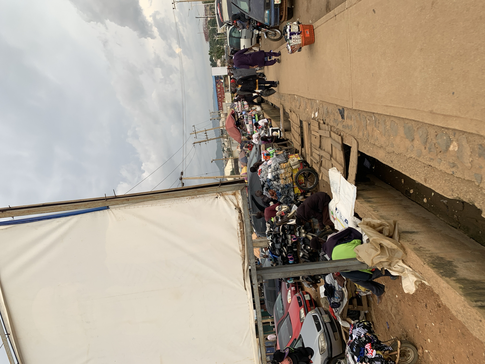 The image depicts a bustling outdoor market scene in Rafin Par, Nigeria. A large white tarp, possibly a stall covering, dominates the left side of the frame. In the foreground, a dirty concrete drain runs along the edge of a dirt path.
Several people are present, engaged in various activities. Some are browsing or selling goods displayed on tables and carts. Shoes and clothing are prominently visible as merchandise. In the background, cars and motorcycles are parked, indicating vehicular traffic. The sky is overcast with clouds, suggesting a cloudy day. The overall impression is one of a vibrant, commercial hub with a mix of pedestrian and vehicular activity.