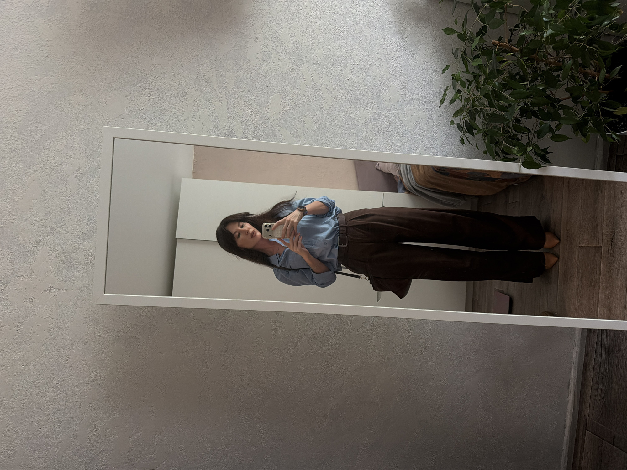 A mirror reflects a woman holding a smartphone, wearing a blue button-down shirt and brown pants. Behind the mirror is a wall with a textured surface and a plant in the upper right corner. The floor appears to be wooden. It is not possible to determine the location in Perm, Russia, from this image.