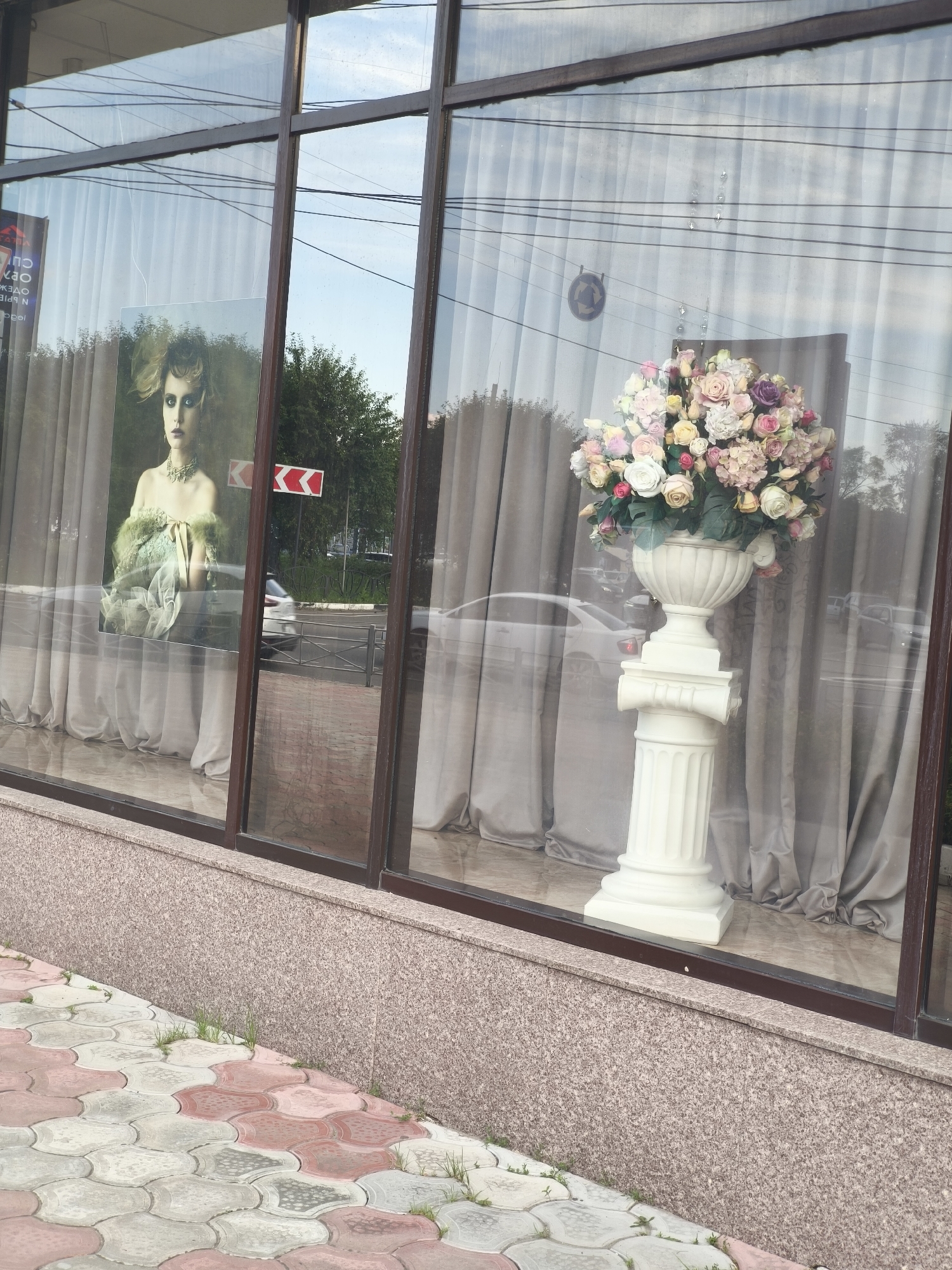 The image shows a storefront window display in Komsomolsk-on-Amur, Russia. The window features a large print of a woman with dramatic makeup and a vintage-style dress. To the right of the print, a large, ornate white vase overflows with a colorful arrangement of artificial flowers. The display is set against a backdrop of beige curtains. Outside the window, a paved walkway with a patterned design is visible, leading towards the street where cars are passing. The building's facade is made of granite. The overall scene suggests a commercial establishment, possibly a boutique or a salon.
