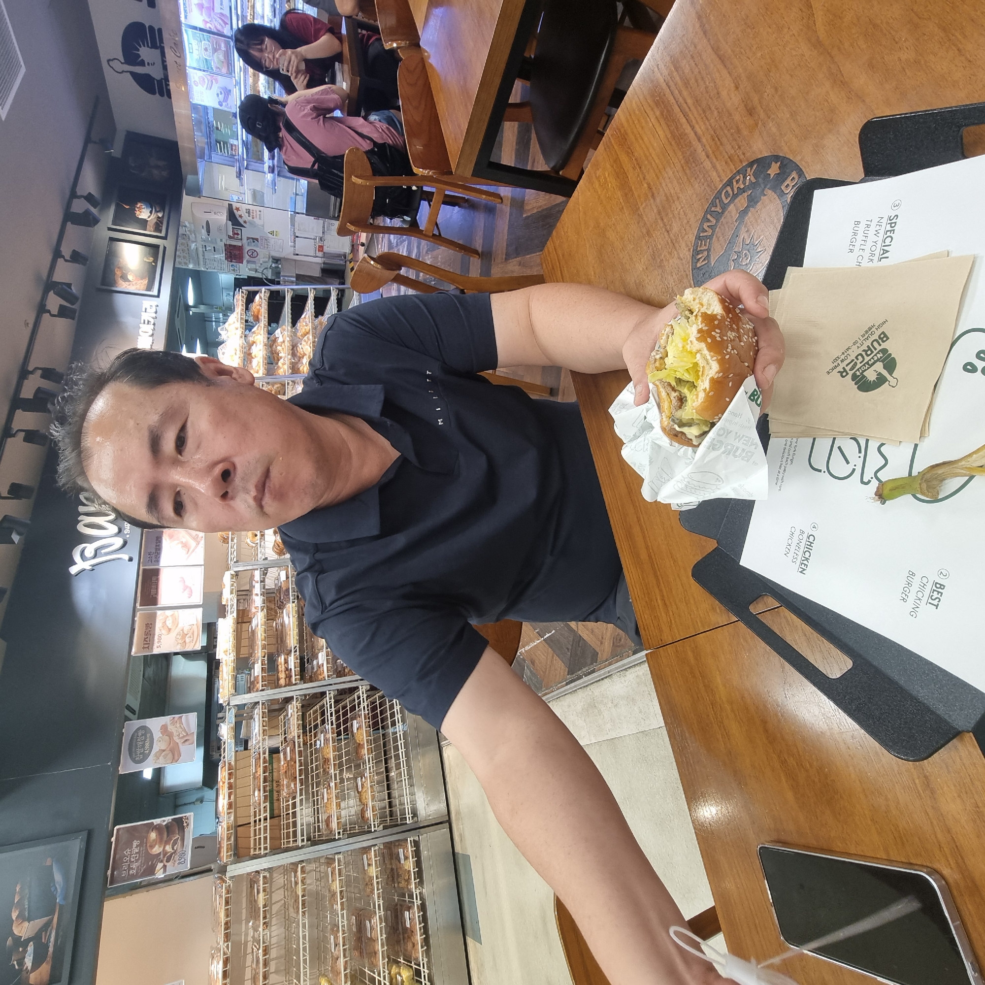 The image depicts an indoor scene at a bakery in Wonju-si, South Korea. A middle-aged East Asian man is seated at a wooden table, holding a half-eaten burger in his right hand. He is wearing a dark blue polo shirt with a small white logo on the left chest. In front of him on the table is a black phone, a white paper wrapper with burger branding, and a menu or placemat. To his right, a circular graphic with the text "NEW YORK BURGER" is visible on the table surface. In the background, shelves filled with baked goods are displayed, and other patrons are seated at tables. One woman with glasses and another with a backpack are visible in the background, along with chairs and tables. The overall environment suggests a casual dining or bakery setting.