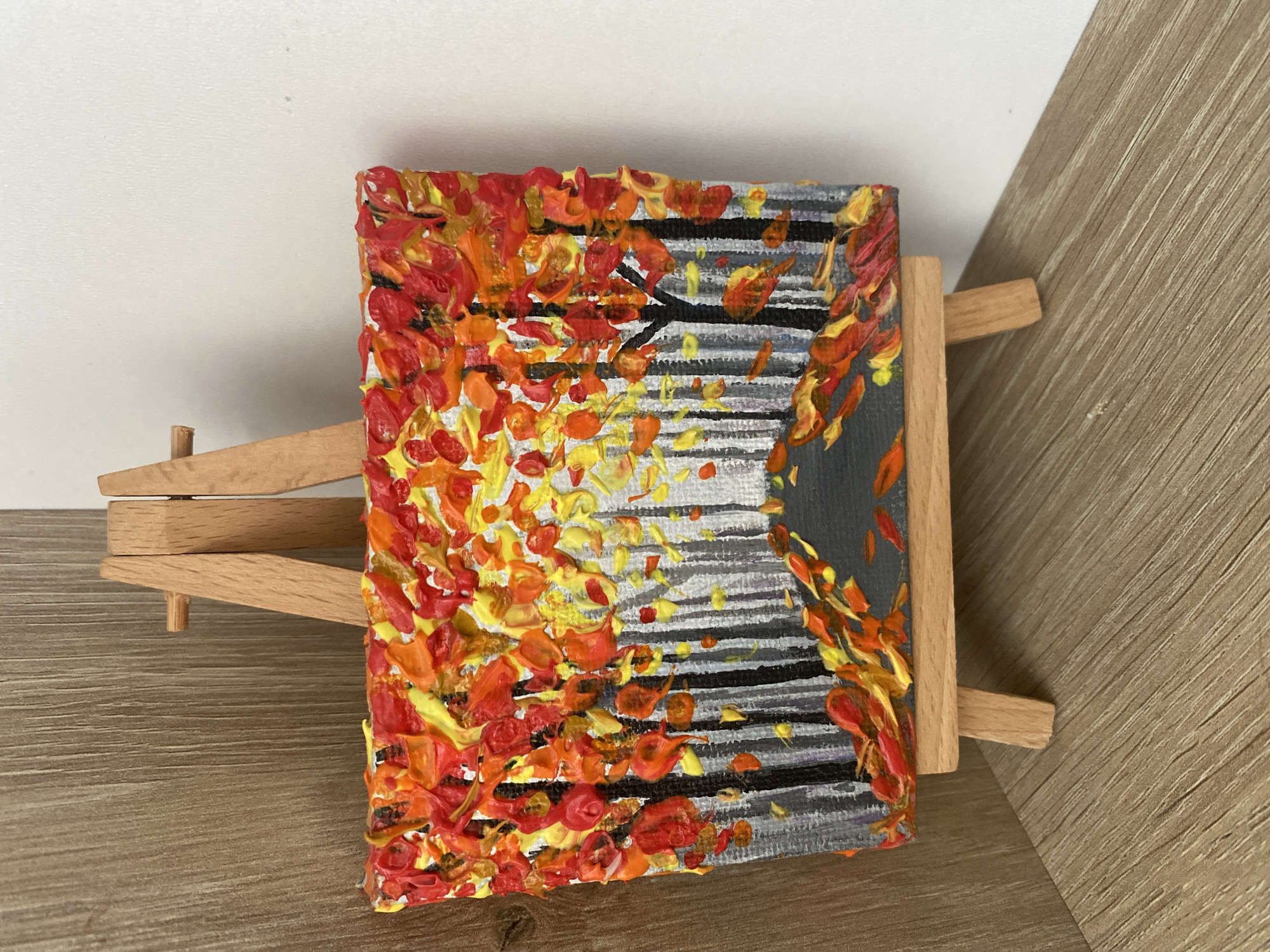 The image shows a small, textured acrylic painting of an autumn forest scene. The painting is displayed on a wooden easel. The foreground depicts a path covered in fallen leaves, painted in vibrant shades of red, orange, and yellow. Tall, thin trees with dark trunks create a sense of depth, and their upper canopies are filled with a dense collection of impasto leaves in similar warm hues, with hints of yellow and bright orange. The background features a slightly blurred, greyish representation of more trees, suggesting a dense forest. The painting's textured surface is evident, with thick applications of paint giving it a three-dimensional quality. The easel is made of light-colored wood and has two supporting legs. The background consists of a wooden surface on the left, with a lighter, plain wall on the right. There is no indication of a specific location in Chișinău, Moldova, or any people or actions within the scene.