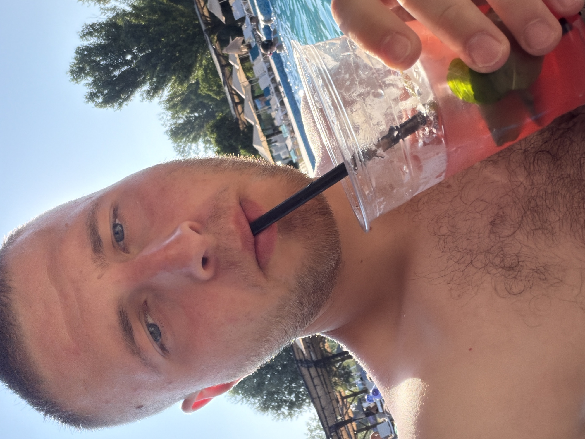 The image is a close-up selfie taken outdoors, likely during the daytime due to the bright sunlight casting shadows and illuminating the scene. The subject is a person holding a clear plastic cup with a red drink, which appears to be a cocktail, with a straw. A few mint leaves are visible in the drink. The person is holding the cup with their right hand, and their arm is visible. The lower part of their face, including their mouth, nose, and eyes, is in the foreground, suggesting the photo was taken from a low angle. The person has short hair and light-colored eyes.
In the background, a swimming pool with blue water is visible, along with lounge chairs and umbrellas, indicating a resort or a recreational area. Green trees can be seen against a clear blue sky, further suggesting an outdoor setting in warm weather. The image is oriented sideways. There is no visible text in the image. The overall mood seems relaxed and summery, implying the person is enjoying a drink by the pool.