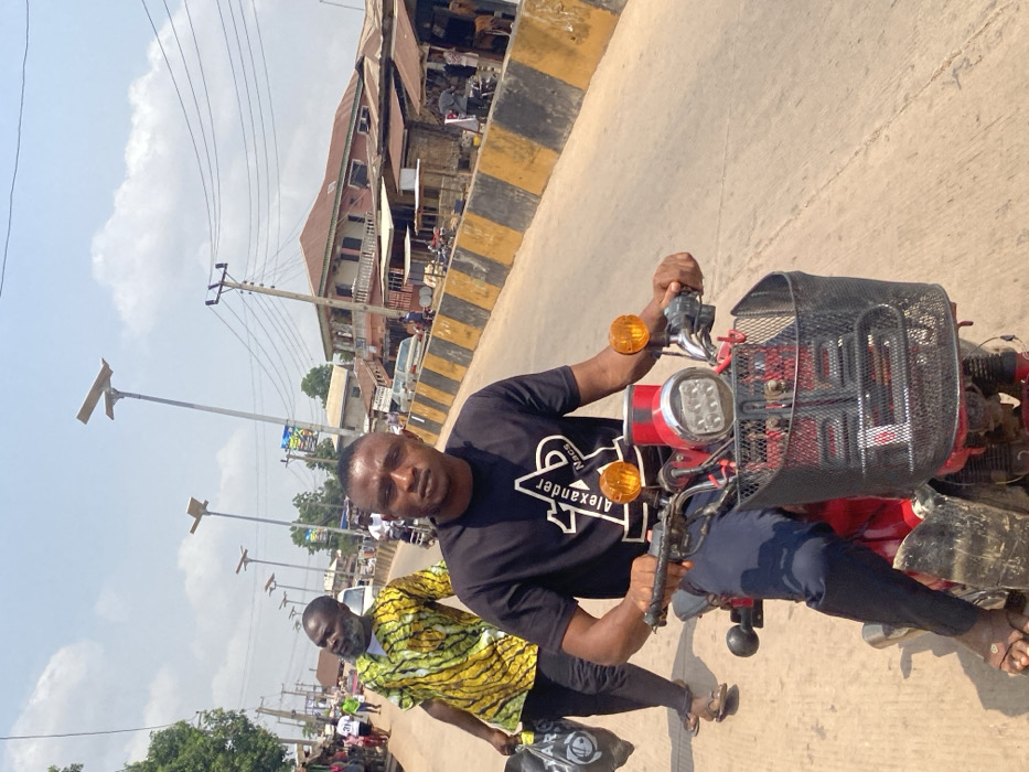 This image depicts a daytime street scene in the city of Afikpo, Nigeria. In the foreground, a dark-skinned male individual, appearing to be in his 20s or 30s, is seated on a red motorcycle, looking directly at the camera. He wears a black t-shirt with "Alexander Macs" branding and dark trousers, with brown sandals on his feet. The motorcycle features a front-mounted black metal basket, a central chrome headlight with multiple LED lights, and two prominent orange turn indicators. His hands rest on the handlebars.
Immediately behind and to the left of the motorcyclist, another dark-skinned male, older with a beard, walks towards the left, carrying a black plastic bag in his left hand. He is dressed in a yellow and green patterned short-sleeved shirt and dark trousers, and is also wearing sandals. His head is tilted upwards and to his left.
The environment includes a paved road surface with a distinct yellow and black striped median or curb running along the right side. On both sides of the road, various buildings are visible, ranging from multi-story structures with balconies to simpler stalls with corrugated metal roofs, suggesting commercial activity. Numerous utility poles with power lines traverse the scene, accompanied by modern, tall streetlights equipped with solar panels. Distant vehicles and pedestrians indicate active street traffic under a bright, partly cloudy sky.