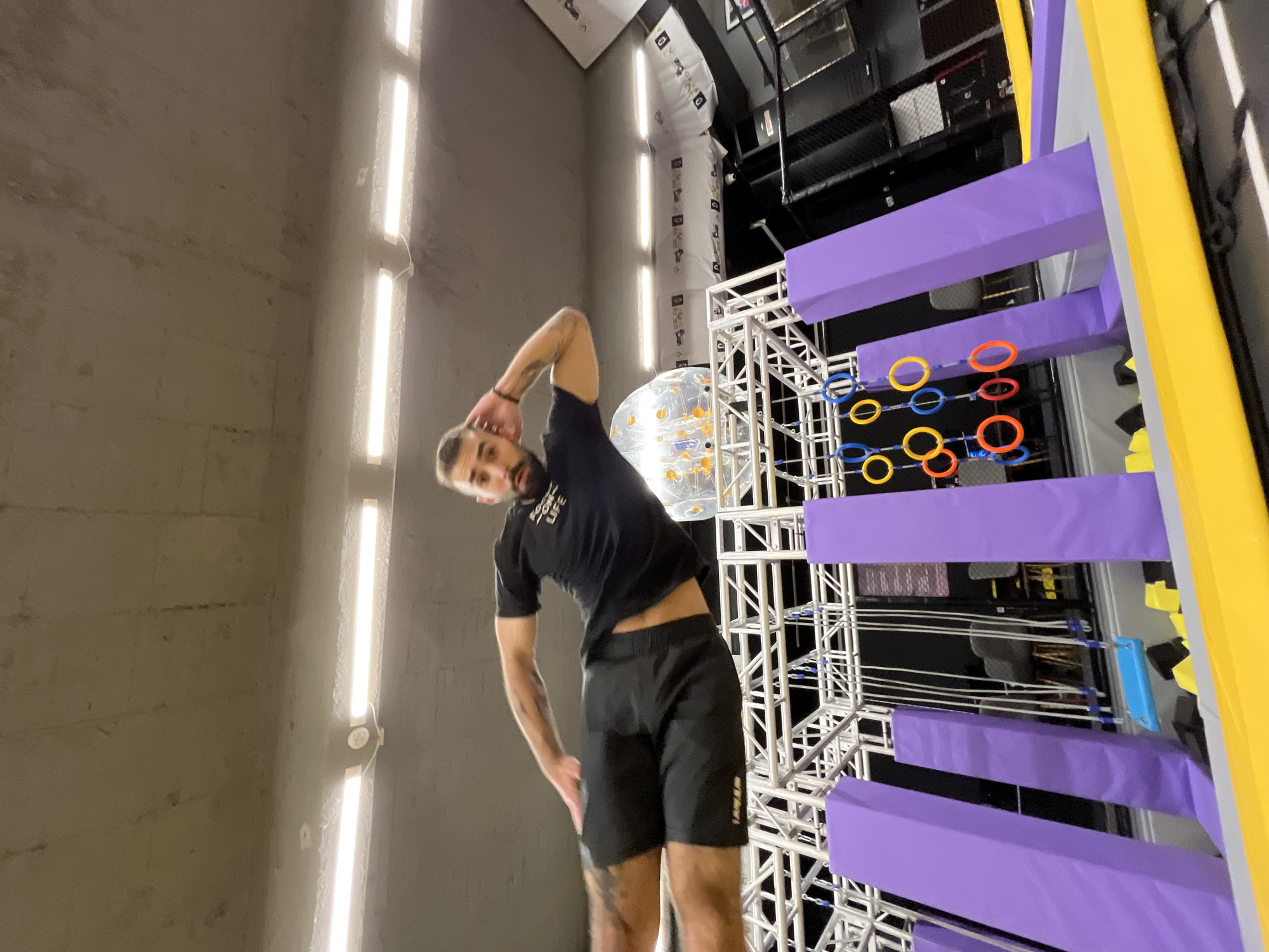 The image depicts an indoor recreational facility, specifically an obstacle course, located in Unknown, Unknown.
In the foreground, a male individual is captured mid-air, positioned horizontally, suggesting a jump or fall. He is fair-skinned with a beard and short dark hair. He wears a black t-shirt with "FOCUS ON LIFE" visible on the front, black shorts, and has tattoos on his left bicep and left thigh. His left arm is bent with the hand near his head, and his right arm is extended forward. His legs are slightly bent.
Below the individual and extending into the mid-ground is a multi-level obstacle course constructed from white metal trussing. This structure supports several purple padded vertical elements. To the right, multiple yellow, blue, and orange hanging rings are visible. Further back and to the left, a blue swing is suspended by ropes. The floor of the obstacle course consists of gray and yellow padded surfaces.
In the background, a large, translucent inflatable sphere with colorful internal elements is visible. The walls are dark, and several white banners or signs with logos and text are affixed to them. An exposed concrete ceiling with multiple linear fluorescent light fixtures illuminates the scene. To the far right, a dark netted enclosure, possibly part of a trampoline or climbing area, is present. Gray upholstered chairs are also visible in the deeper background.