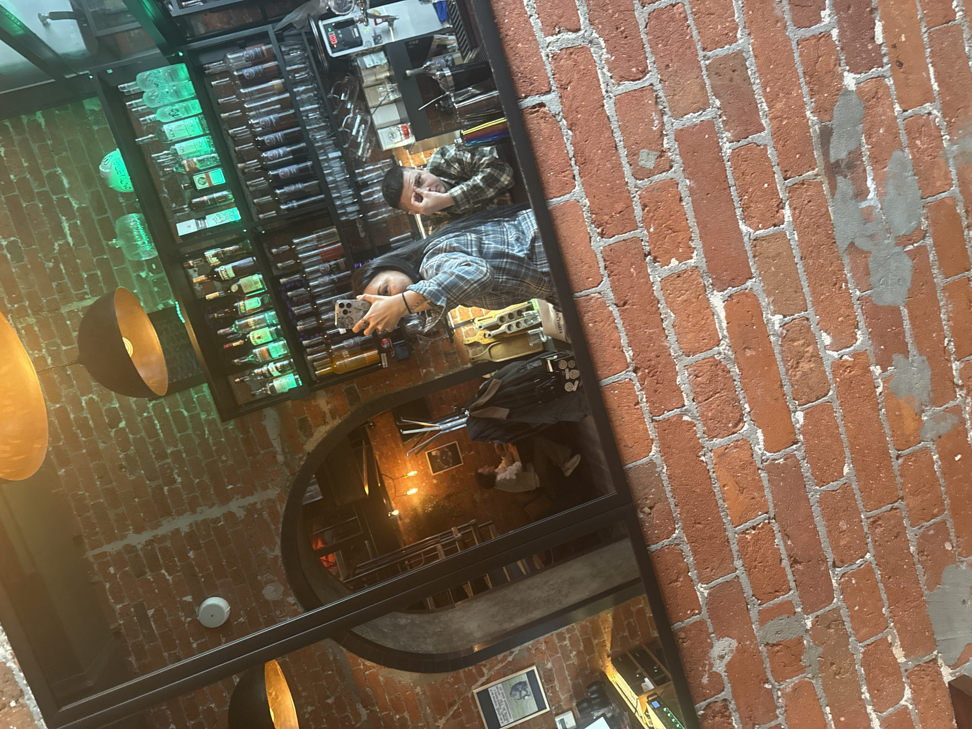 The image is a reflection in a mirror of a bar or restaurant interior, showing a couple taking a selfie. The man and woman are wearing plaid shirts and are in the upper portion of the reflection. The woman is holding a smartphone and taking a picture. In the background, there is a bar shelf stocked with alcohol bottles, illuminated with green light. The room also features brick walls, and there are hanging lights in the foreground. The mirror’s frame is visible, along with portions of brick wall on either side.