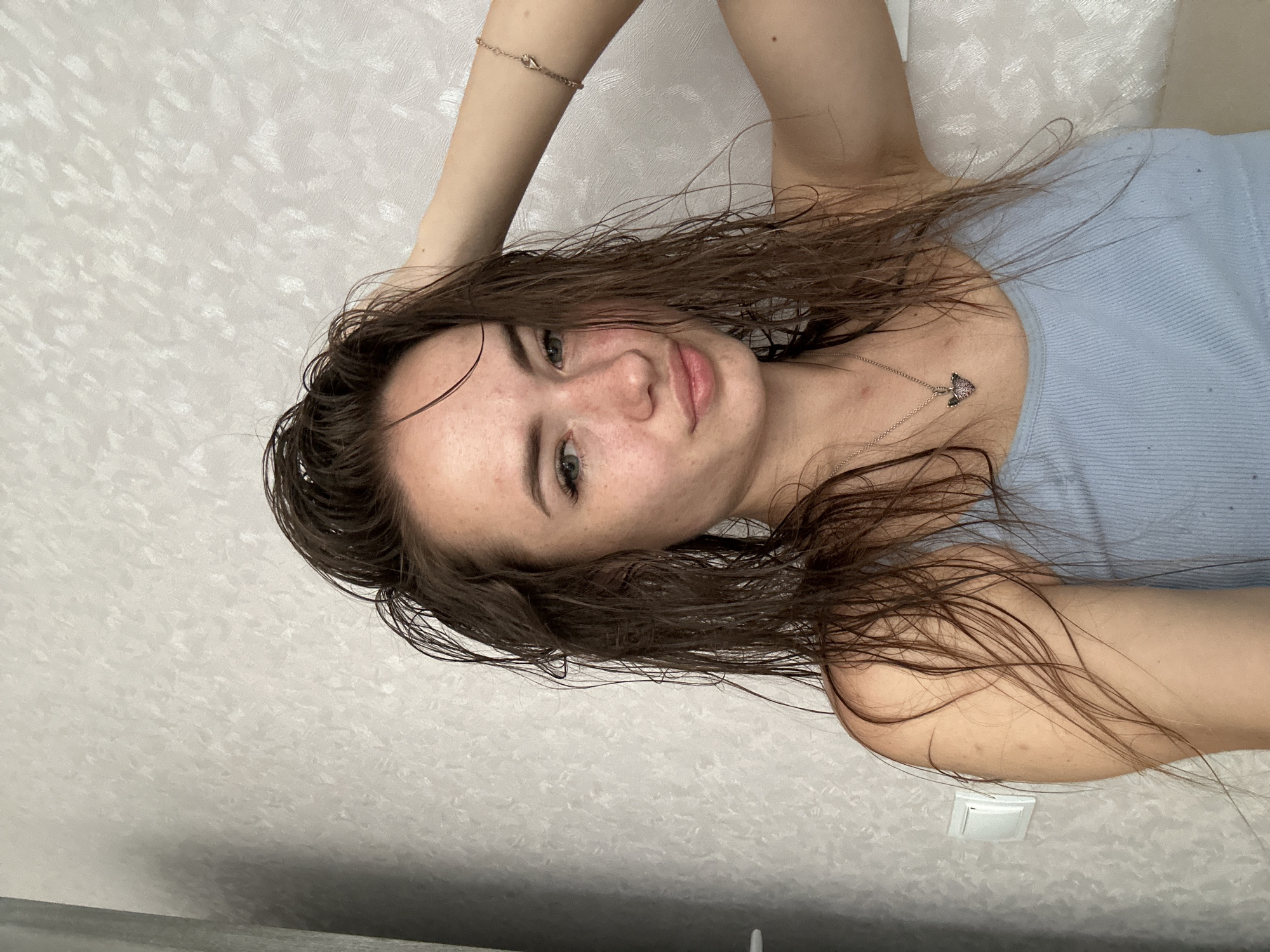 A young woman with wet brown hair is shown from the chest up, looking at the camera. She has freckles across her nose and cheeks and is wearing a light blue tank top. Her right hand is resting on her head, and she is wearing a bracelet on her right wrist and a necklace with a pendant. The background is a textured, light-colored wall. The scene appears to be indoors, with a light switch visible on the left side of the image.