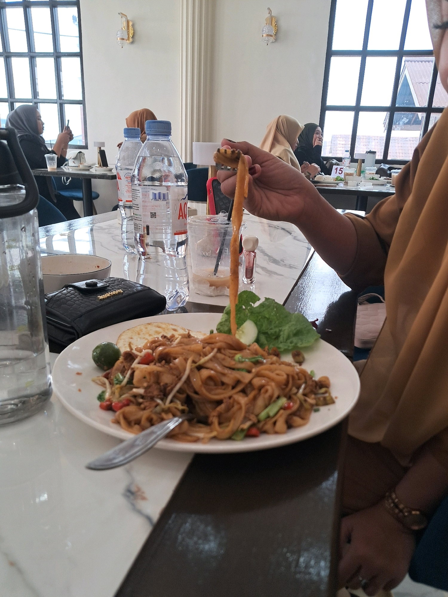 The image captures a close-up of a meal on a white plate placed on a marble-patterned table. The meal consists of noodles with what appears to be vegetables and meat, alongside a fried egg and a small lime. A fork with noodles is being held up, mid-air, by a person whose hand and arm, wearing a light brown hijab, are visible in the foreground. To the left of the plate, a transparent water bottle is partially visible. Behind the meal, two bottles of water are prominently displayed. In the background, several people are seated at tables, some wearing hijabs. The setting appears to be an indoor restaurant with large windows offering a view of a building's roof. Based on the context of Mempawah, Indonesia, this scene likely depicts a meal at a local eatery.