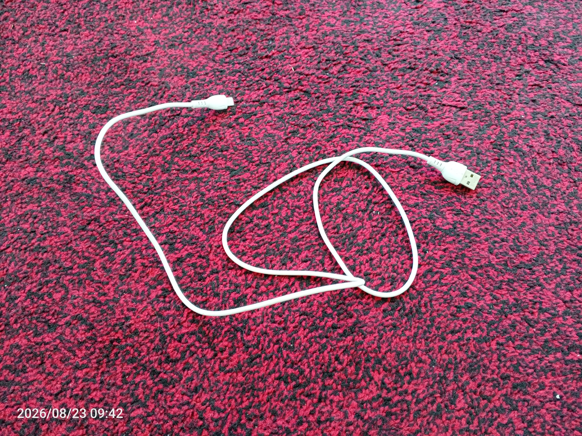 A white USB-C cable is laid out on a red and black textured carpet. The cable has two distinct ends, one appearing to be a USB-A connector and the other a USB-C connector. The USB-A connector is visible on the right side of the image, while the USB-C connector is on the left. The cable forms a loose loop in the center of the frame. The carpet has a dense pile with alternating red and black fibers, creating a speckled pattern. The timestamp "2026/08/23 09:42" is present in the bottom left corner of the image. The scene is indoors, on a carpeted surface. No people or animals are present in the image.