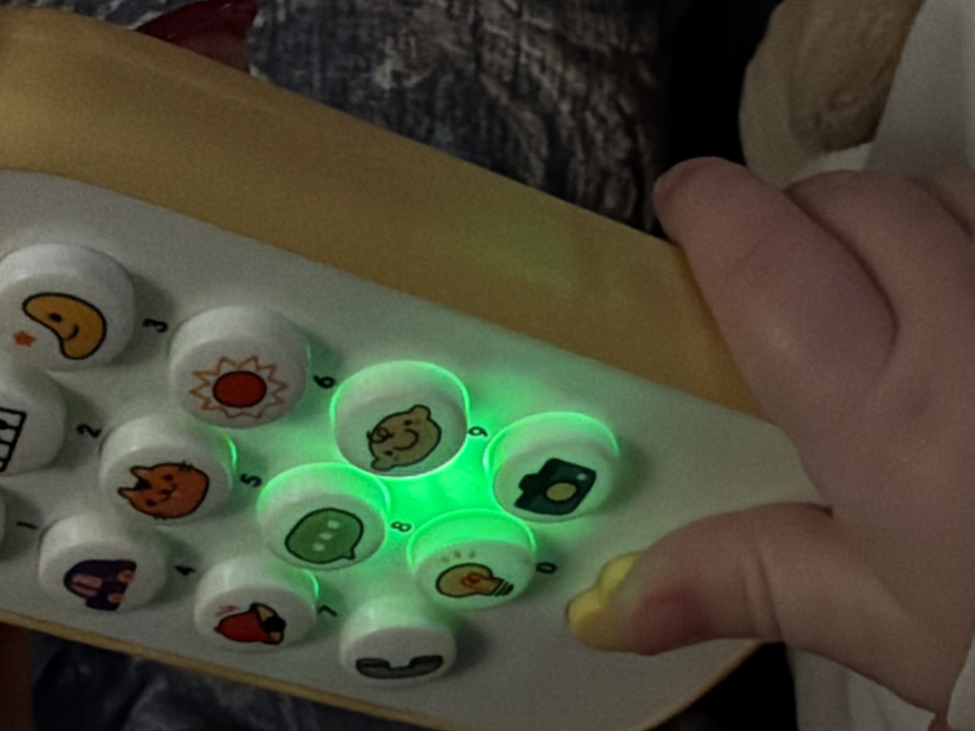 A close-up shot shows a child's hand interacting with a toy phone. The phone is white with a yellow border and features circular buttons with various icons such as a sun, cat, speech bubble, lightbulb, and camera. Several of the buttons are illuminated with a green light. The child's hand is positioned to the right of the phone, with a thumb resting near the bottom right corner. The background is partially visible and shows dark fabric, and what appears to be a white shirt. Location details of Yerevan, Armenia are not possible to determine from this single image.