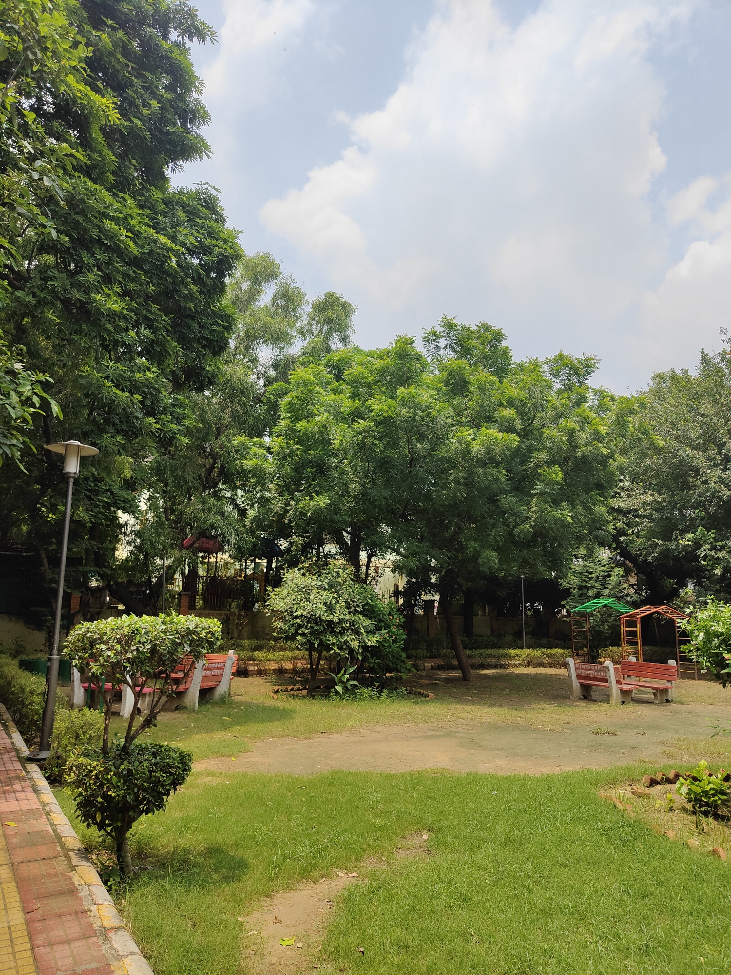 The image shows a park scene in Gurgaon, India. In the foreground, a paved pathway made of yellow and red bricks runs along the left side. Adjacent to the pathway is a well-maintained green lawn with a small, intricately shaped bush in the center. Further into the park, there are several benches, some with red seating and concrete bases. A lamppost stands near the pathway. Lush green trees of various sizes dominate the background, providing ample shade. In the middle ground, there are more benches and a covered seating area with a red roof. The sky above is partly cloudy with patches of blue. The overall impression is a serene and verdant public space.