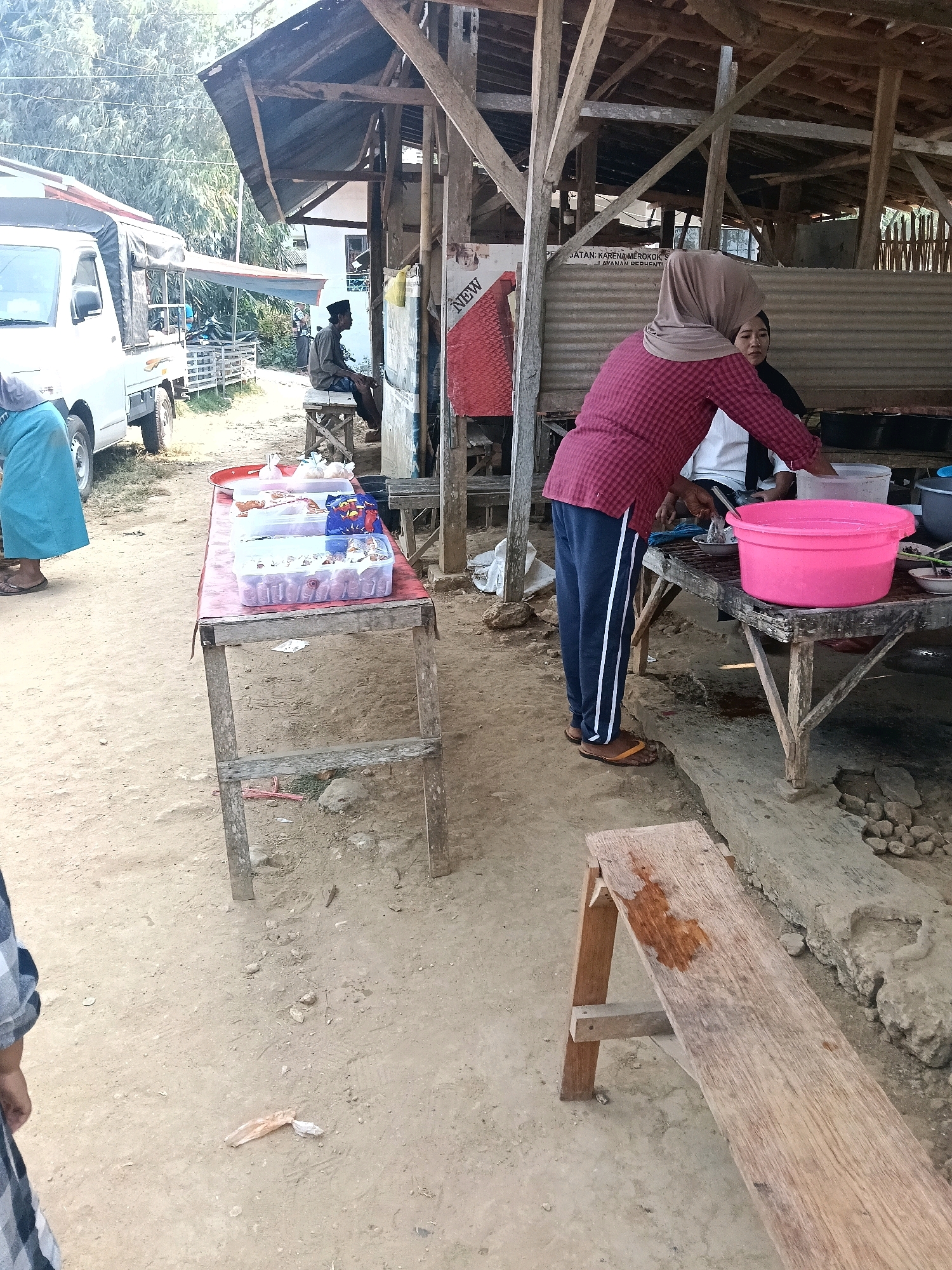 A woman wearing a maroon checkered shirt and navy blue pants with white stripes, along with a hijab, is standing and preparing food at an outdoor food stall in Tebul Barat, Indonesia. She is standing behind a table displaying various food items in clear plastic containers. To her right, another woman in a white shirt and black hijab is seated, looking at the food. In the foreground, a long wooden bench is placed on the dirt ground. A white truck is visible on the left side of the image, parked near the food stall. The stall appears to be constructed from wood and corrugated metal, with a rustic, open-air design. The ground is dusty and uneven, with scattered rocks. Trees and foliage are visible in the background.