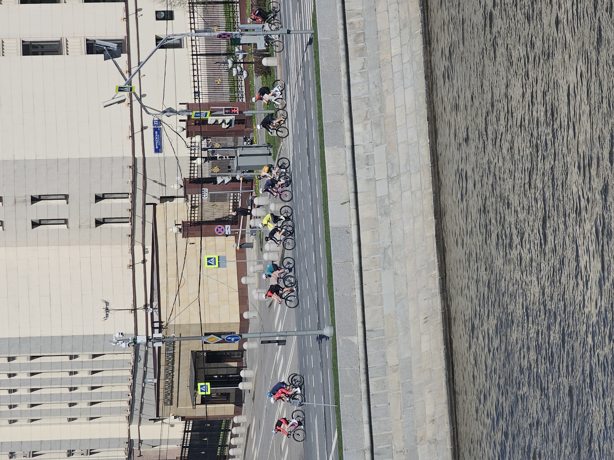 The image shows an aerial view of a group of cyclists riding along a street in Moscow, Russia. The street runs alongside a river with a stone embankment. On the left side of the street is a tall building with a beige and gray facade. Several traffic signs and street lights are visible along the street. The cyclists are dressed in various athletic attire. The street appears to be well-maintained with lane markings.