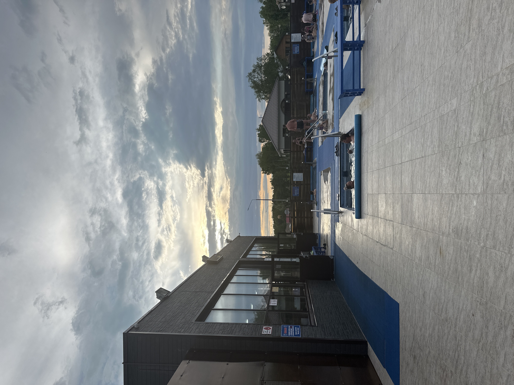 In Yuzhnyye Koryaki, Russia, an outdoor recreational facility features a pool area with several small, tiled pools, some with blue decking and benches. Several individuals are present in and around the pools, some bathing and others standing or sitting. A dark-colored building with large windows, possibly a changing room or cafe, is situated to the left of the scene, with a blue mat extending from its entrance towards the pool area. The sky above is overcast with dramatic clouds, and the sun is setting, casting a warm glow on the horizon. The overall environment appears to be a public leisure space.