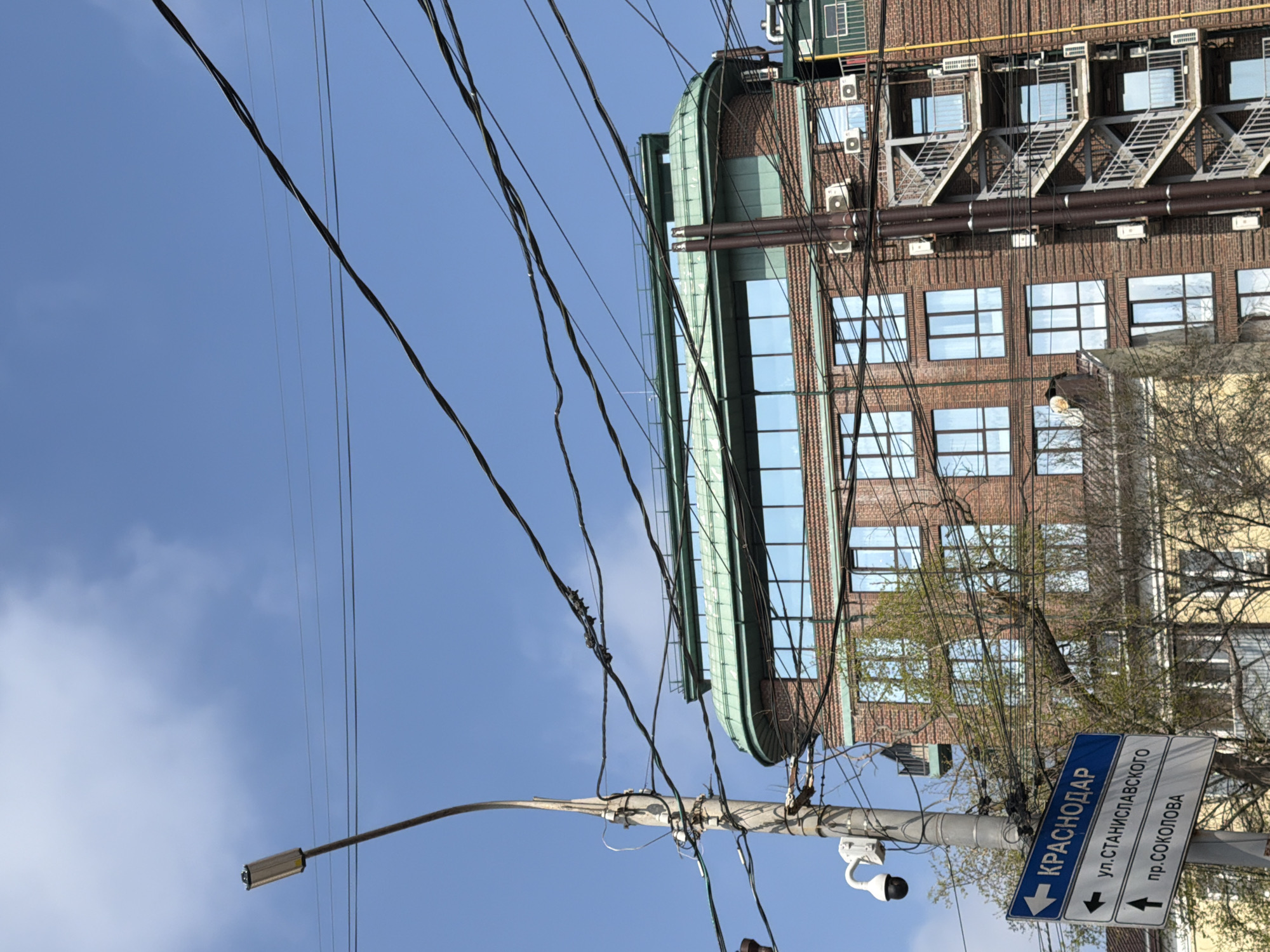 The image shows a street scene in Krasnodar, Russia, likely taken from a low angle. A brick building with numerous windows, some reflecting the sky, is visible. It also has fire escapes and various pipes/vents running along its facade. In the foreground, there's a street sign indicating "Krasnodar" and street names "ул. Станиславского" and "пр. Соколова." A utility pole is present, adorned with numerous electrical wires and a streetlight. A security camera is attached to the pole. The sky is blue with some scattered clouds.