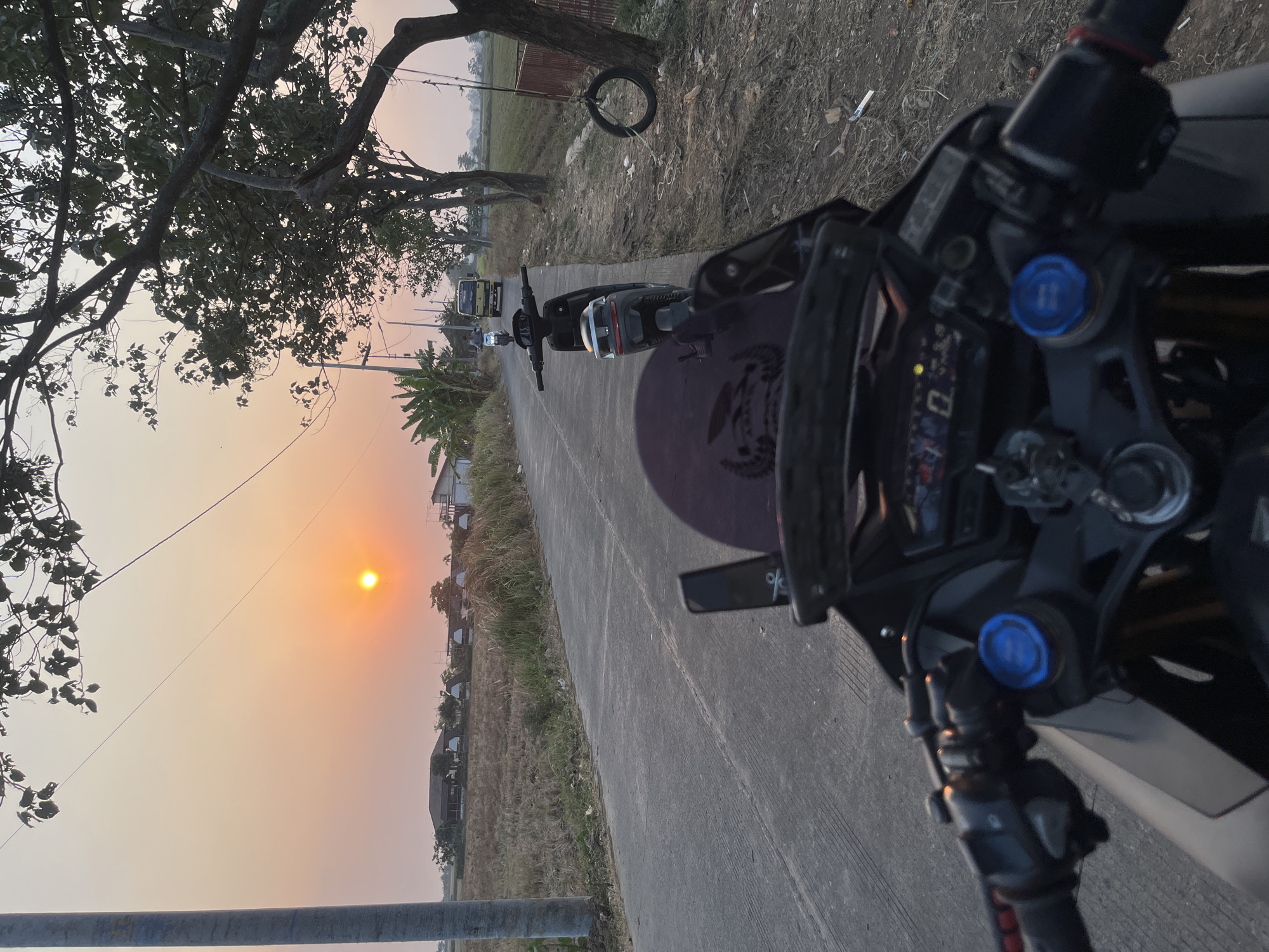 The image depicts a scene on a rural road in Dusun Dua, Indonesia, during sunset. A motorcycle, viewed from the rider's perspective, is parked on the side of the road. The motorcycle has a black body, a tinted windscreen, and a digital dashboard. In the background, another motorcycle and a yellow truck are visible on the road. The landscape features dry grass fields, trees, and a few houses. A tire swing hangs from a tree on the right side of the image. The sky is illuminated by the warm glow of the setting sun. The overall atmosphere is serene and captures a moment of rural life.