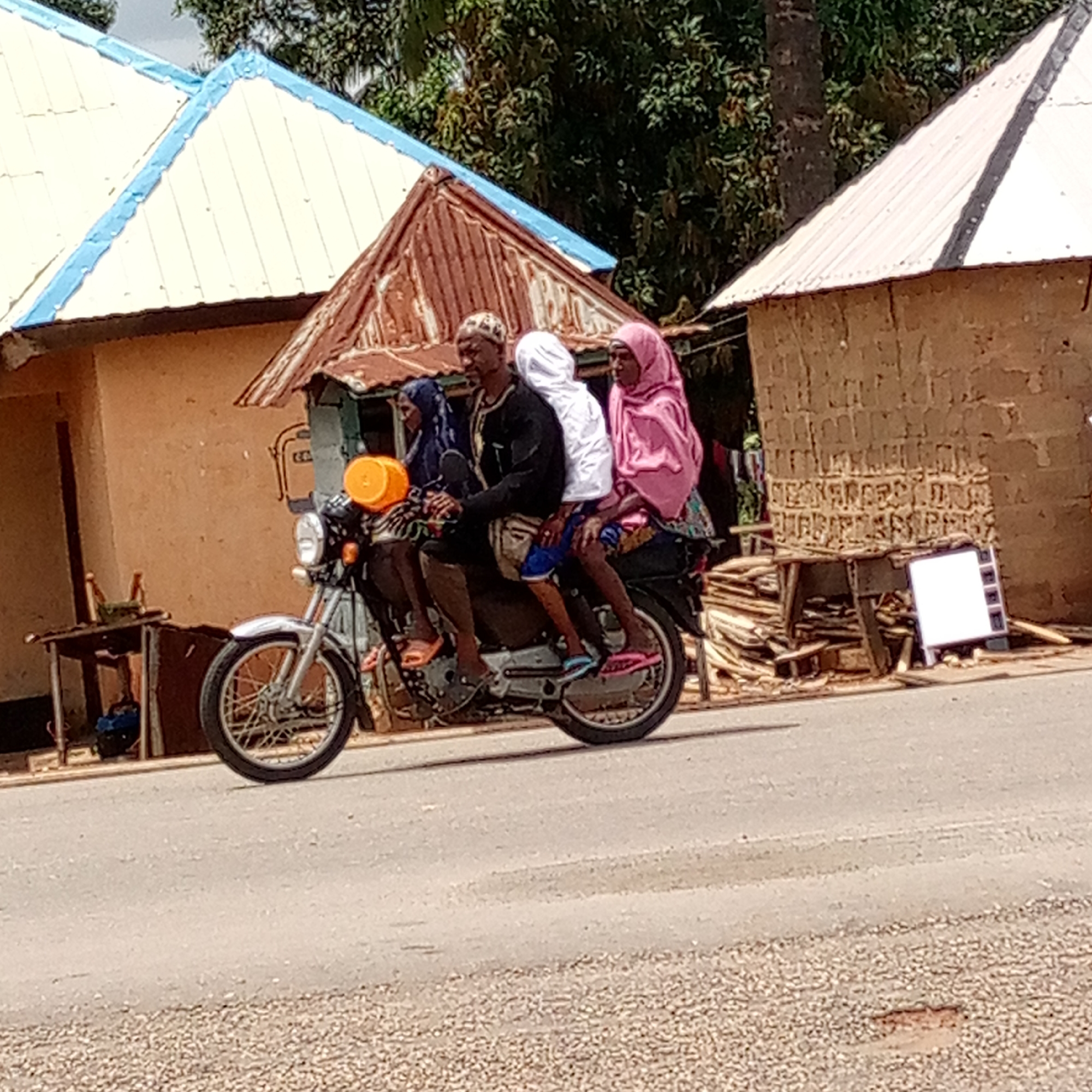 The image depicts a motorcycle carrying four individuals, moving on a paved road in Mararaba, Nigeria. The rider is a man wearing a black shirt, patterned head covering, and shorts. In front of him, a woman in a blue hijab is seated, and behind him are two other passengers: a person in white and a woman wearing a pink hijab. The motorcycle has an orange bucket strapped to the front. In the background, there are buildings constructed of clay bricks and corrugated metal roofs, and there are several wooden tables and piles of lumber. The road is paved with asphalt and there is gravel on the sides.