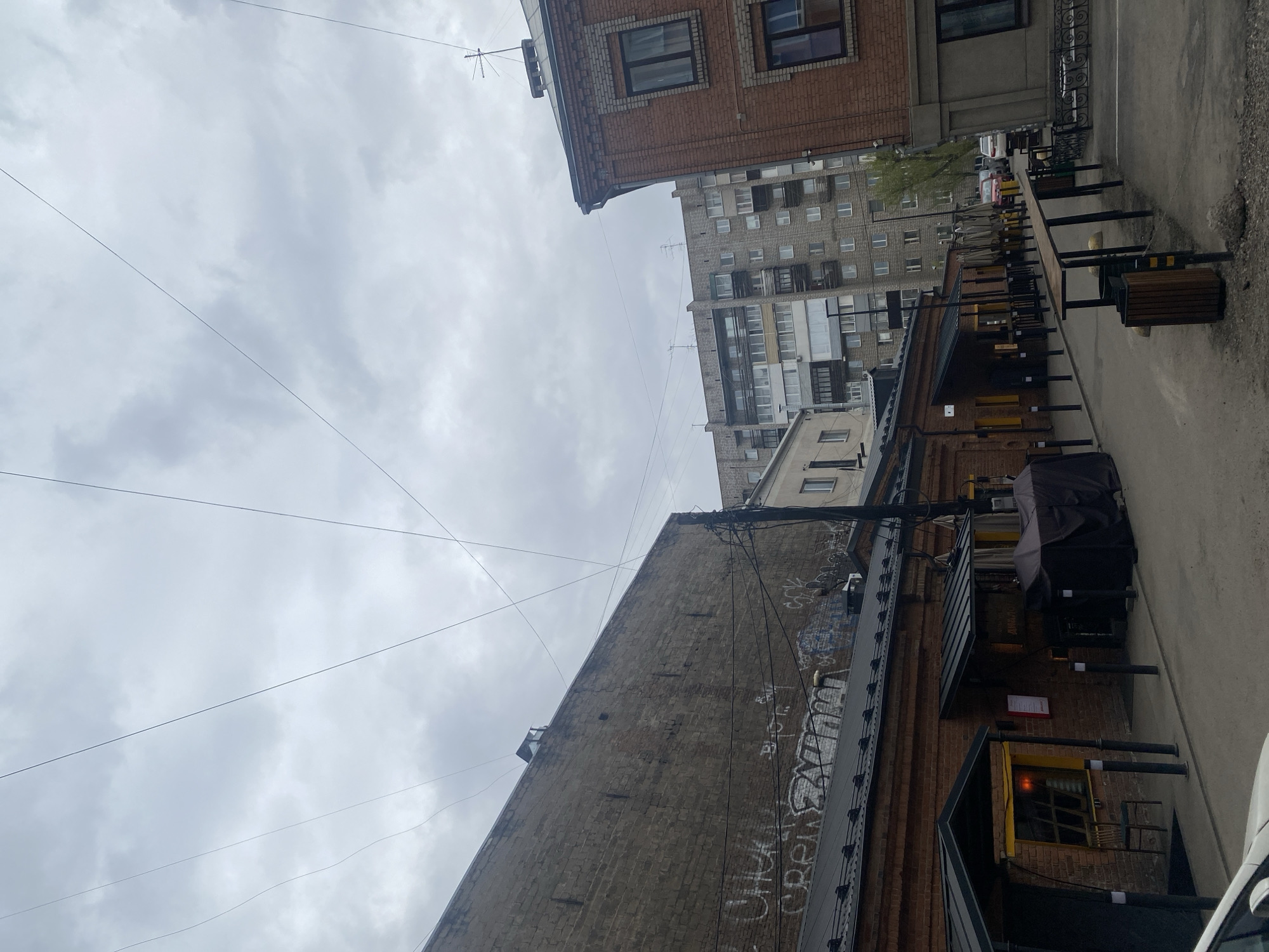 The image depicts an urban alleyway in Krasnoyarsk, Russia. Tall buildings frame the narrow street. The building on the left shows signs of age, with exposed brick and graffiti along its walls. Overhead, a network of utility lines cuts across the sky, which is overcast. The alleyway features a covered outdoor dining area with metal frames and dark umbrellas. One can notice a white vehicle and some outdoor seating arrangements along the side.