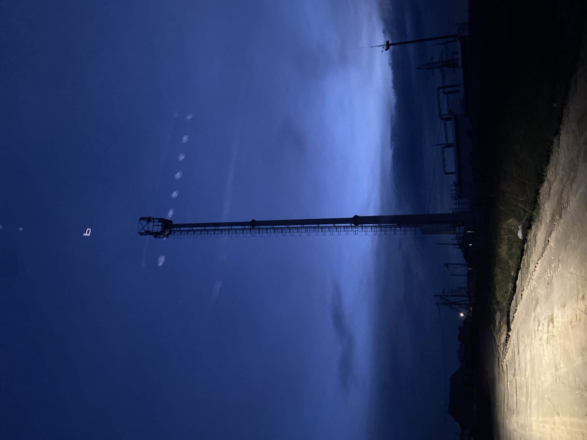 The image, oriented correctly, depicts a nocturnal or late twilight scene dominated by a tall, slender metal lattice tower standing prominently against a dark blue, cloudy sky. The tower, possibly a communication or observation mast, features a platform at its top.
The sky is a deep, rich blue, with cloud formations visible, particularly towards the right side where the clouds appear lighter. Several faint, circular light artifacts, possibly lens flares or reflections, are scattered diagonally across the upper-left sky, alongside a small, light-colored object resembling an inverted "n" or "u."
To the right of the tower, stretching across the middle ground, lies a dark, silhouetted industrial landscape. This area is characterized by numerous power lines, utility poles, and what appear to be structures belonging to an electrical substation or a similar industrial facility.
In the foreground, on the right side, an unpaved road, likely dirt or gravel, is brightly illuminated by an artificial light source, strongly suggesting a vehicle's headlight. The light casts a yellowish glow on the uneven road surface. Sparse vegetation or grass lines the edge of the road and appears in the darker industrial areas.
The scene conveys an atmosphere of an industrial or infrastructure-heavy area, potentially on the outskirts of Chaikovsky, Russia, captured during low light conditions. No specific human activity is visible, and there is no discernible text.