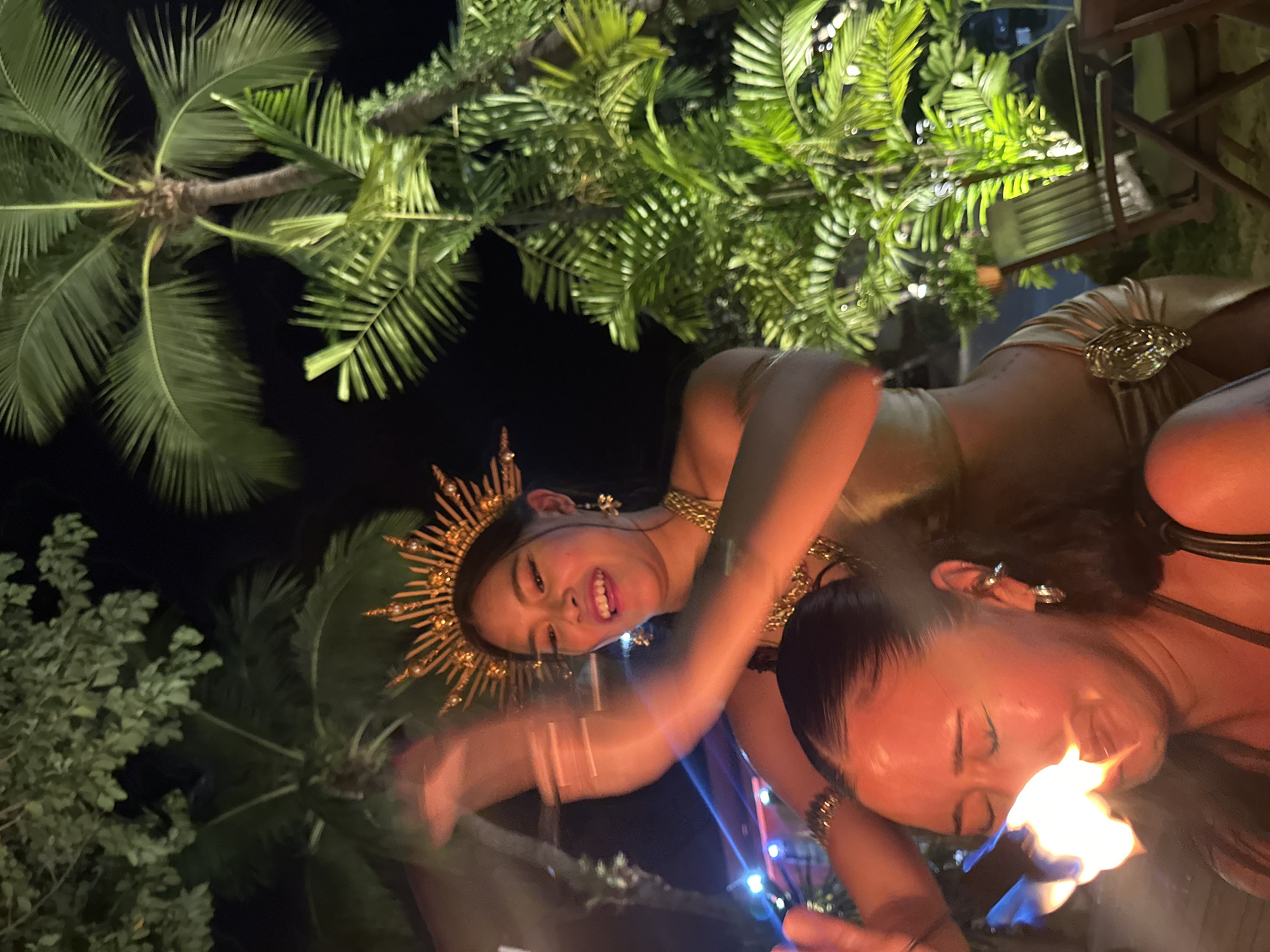 Two women are performing outdoors at night, likely in Baan Chaweng, Thailand, given the presence of palm trees. One woman is wearing a golden, sun-like headdress and a gold-colored outfit, smiling and facing the camera. The second woman is holding fire near her mouth. Both women appear to be adorned with gold jewelry. The surrounding environment includes foliage, including palm trees. Artificial lighting is also present.