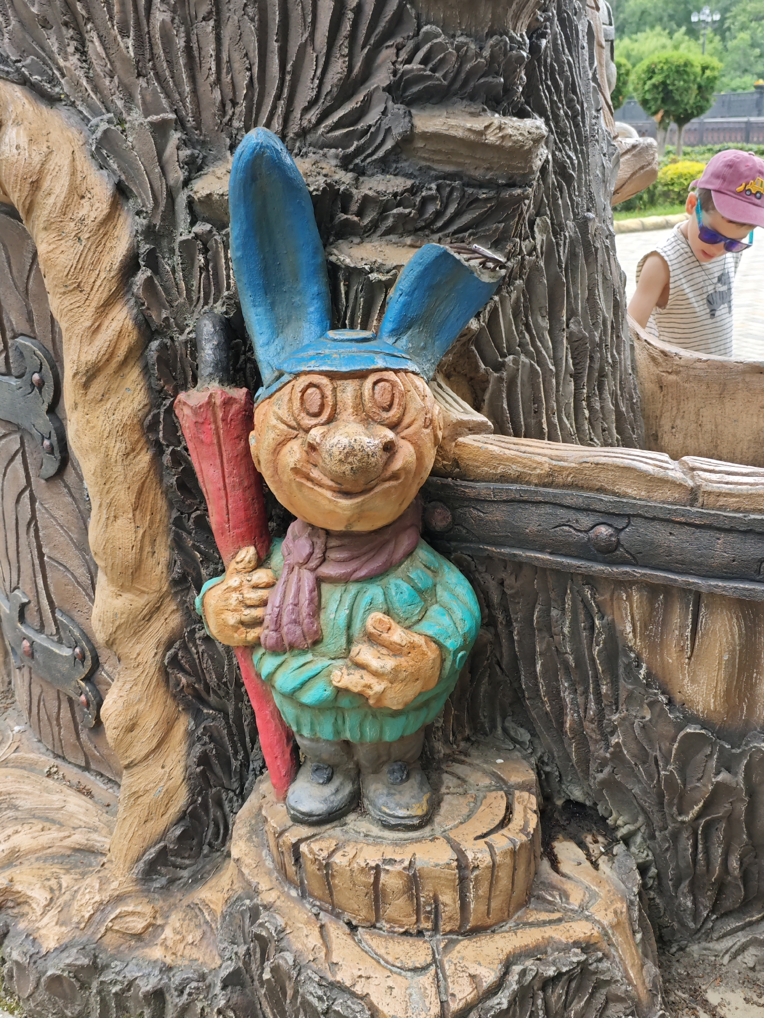 This is a full-shot, eye-level photograph of a garden gnome statue situated in front of a textured, tree-like sculpture. The gnome, made of what appears to be concrete or plaster, is depicted with large blue rabbit ears, a blue cap, and a friendly, slightly mischievous expression. It is wearing a teal tunic, a purple scarf, and black boots. In its right hand, it holds a red object resembling a rolled-up scroll or a flag. The gnome stands on a log-like base, which is part of the larger sculpture.
In the background, a young boy wearing a striped shirt, a pink baseball cap with a yellow construction vehicle emblem, and blue sunglasses, is partially visible, looking downwards as he interacts with the sculpture. The environment suggests an outdoor setting, possibly a park or a playground, with greenery and trees visible in the far background. The overall aesthetic of the sculpture and the presence of the child suggest a whimsical or storybook-themed area.
This scene is located at the Cherkessk City Park of Culture and Leisure in Cherkessk, Russia, specifically within a decorative area featuring fairy tale characters and installations.