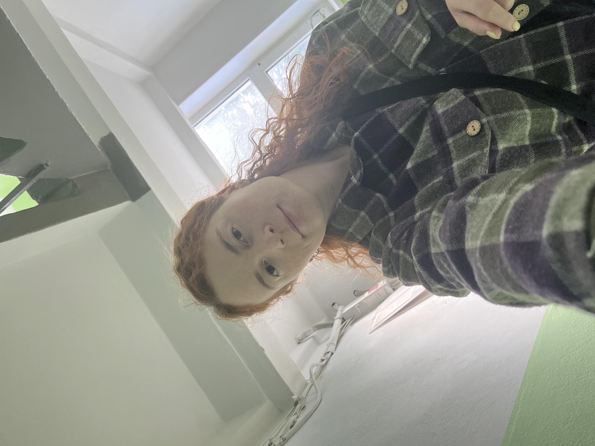 A selfie image showing a woman with red curly hair wearing a green and black plaid shirt. The image appears to have been taken in the stairwell of a building. The background is primarily white with some green sections near the bottom right corner. A window is visible in the top center of the image. Electrical wiring runs along the wall. The photograph was taken in Chaikovsky, Russia.