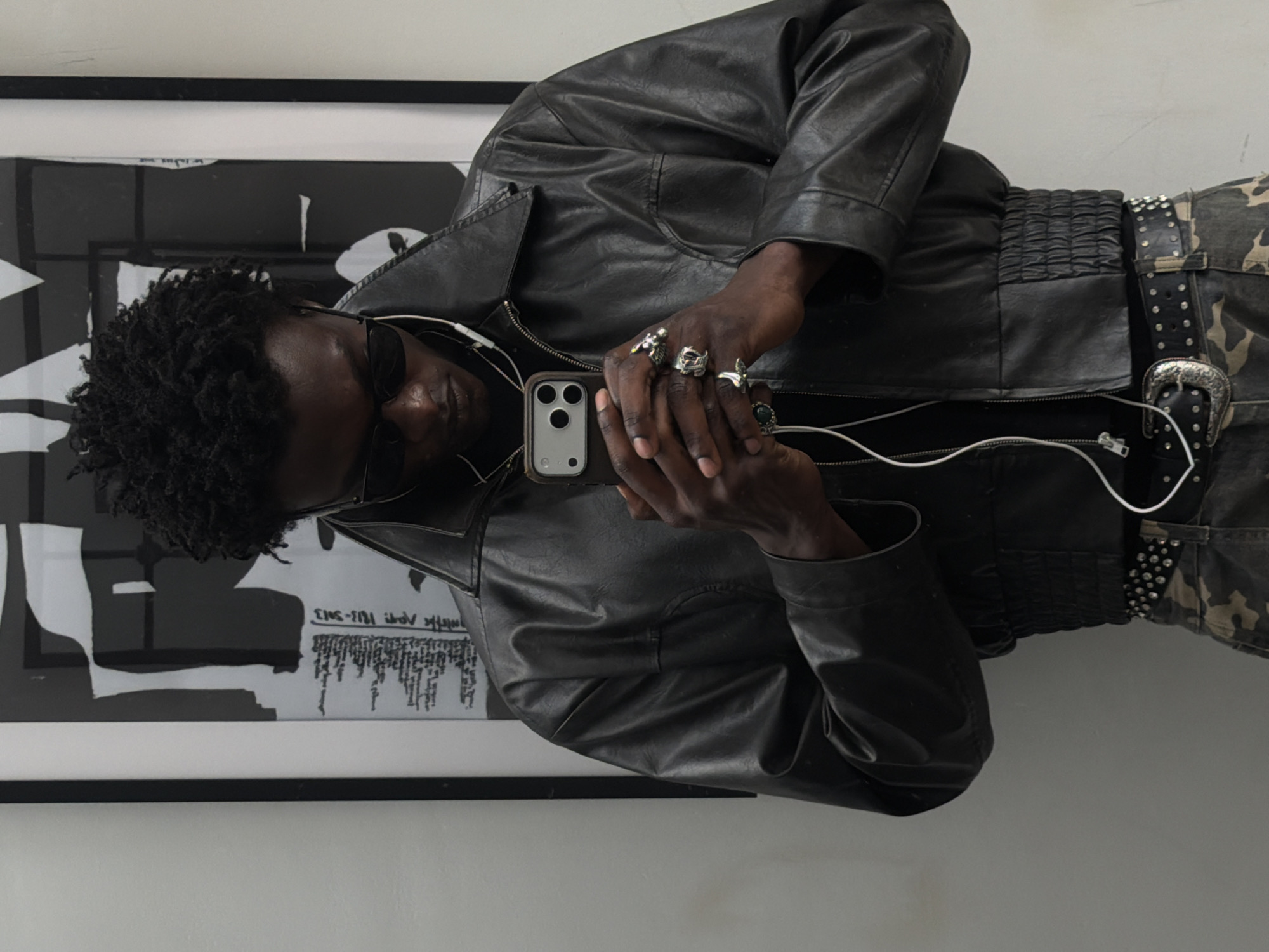 A man with dark skin and dark curly hair, wearing sunglasses and a black leather jacket with a black shirt underneath, is holding a smartphone. He has multiple rings on his fingers. He is wearing a studded black belt and camo pants. The background includes a framed piece of artwork with black and white abstract shapes and text. The image is taken indoors against a neutral colored wall. It is unclear if the location is in London, United Kingdom.