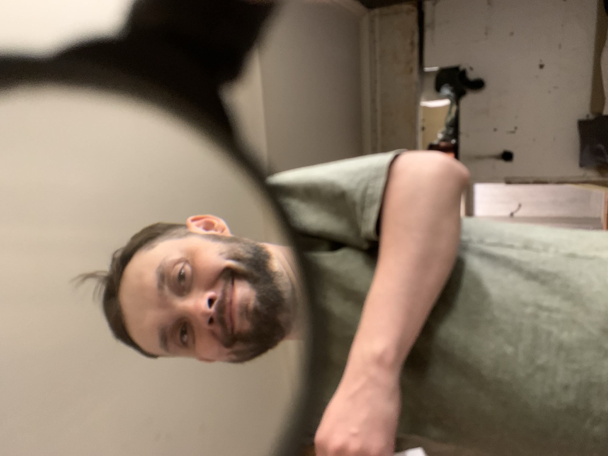 A man is reflected in a handheld round mirror, seemingly taking a selfie. He has brown hair, a beard, and is wearing a green t-shirt. The background is blurry, but appears to be an indoor space with white walls. A metal object is visible in the background. The location is unknown; the city of Zasechnyi, Russia cannot be confirmed.