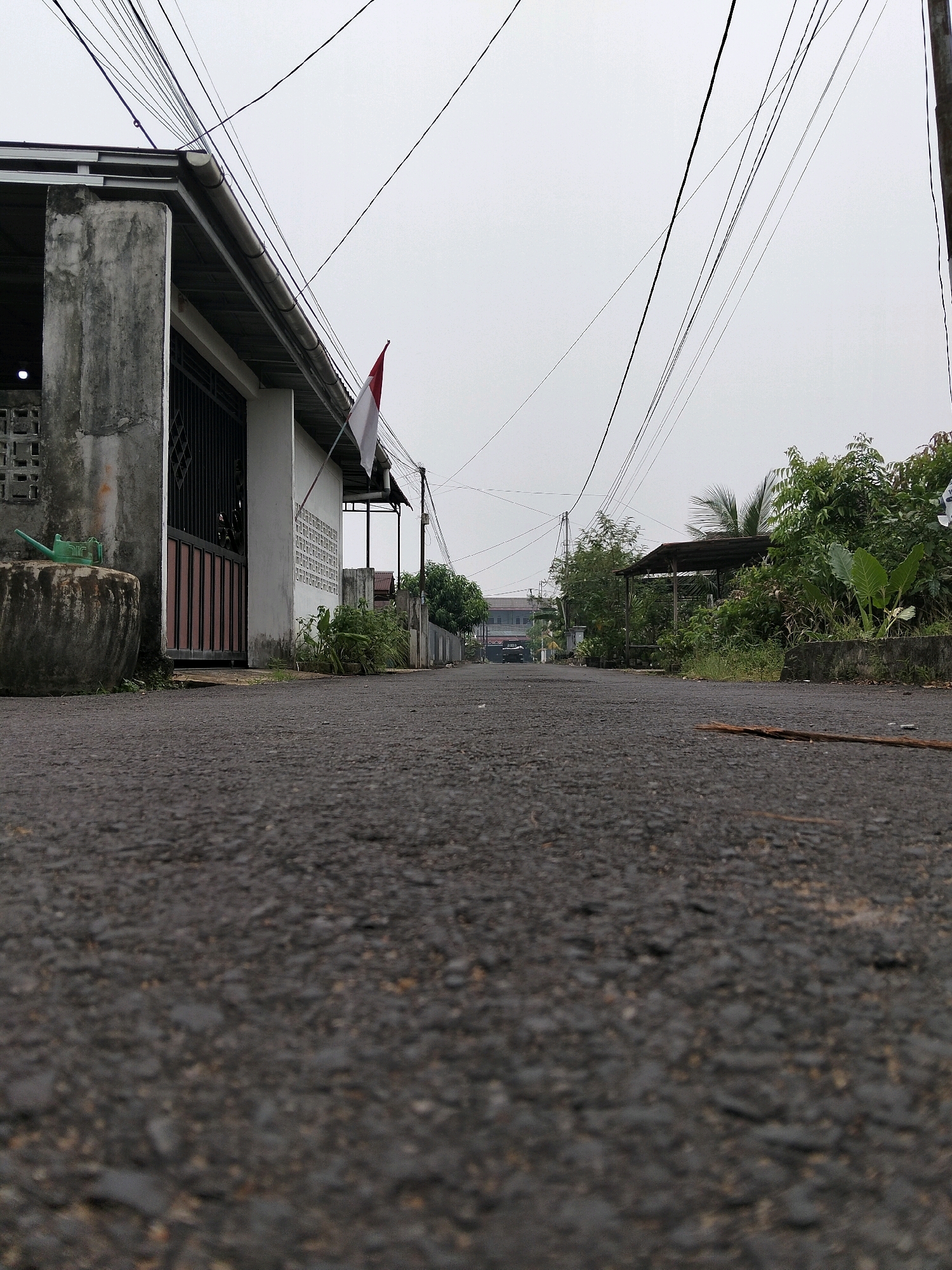 The image depicts a low-angle view of an asphalt road in Arang Limbung, Indonesia, on an overcast day. To the left, a weathered concrete structure with a white wall and a metal gate is visible. A red and white flag, likely the Indonesian flag, is attached to the building. A large concrete pot with a green watering can sits in front of the building. The road recedes into the distance, flanked by vegetation and more buildings. Numerous electrical wires crisscross the sky above. The overall impression is one of a quiet, possibly residential, street.