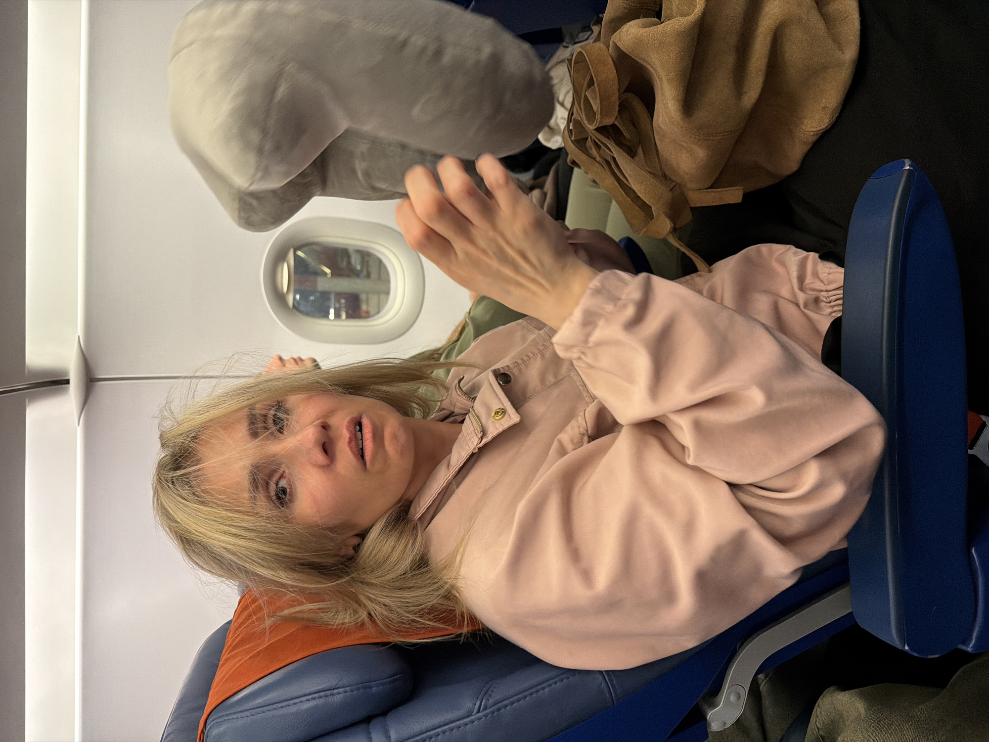 A young adult woman with blonde hair is sitting on an airplane, looking distressed. She is wearing a light pink jacket and her hands are raised in front of her face. She is seated next to a window, and a grey travel pillow is visible on her right. A brown suede bag is on her lap. The interior of the airplane is visible, with blue seats and white walls. The woman appears to be in distress, with her mouth open and her eyes wide. The overall scene is dimly lit, with the primary light source coming from the airplane's interior lights. The location of Saint Petersburg, Russia is not depicted in the image.