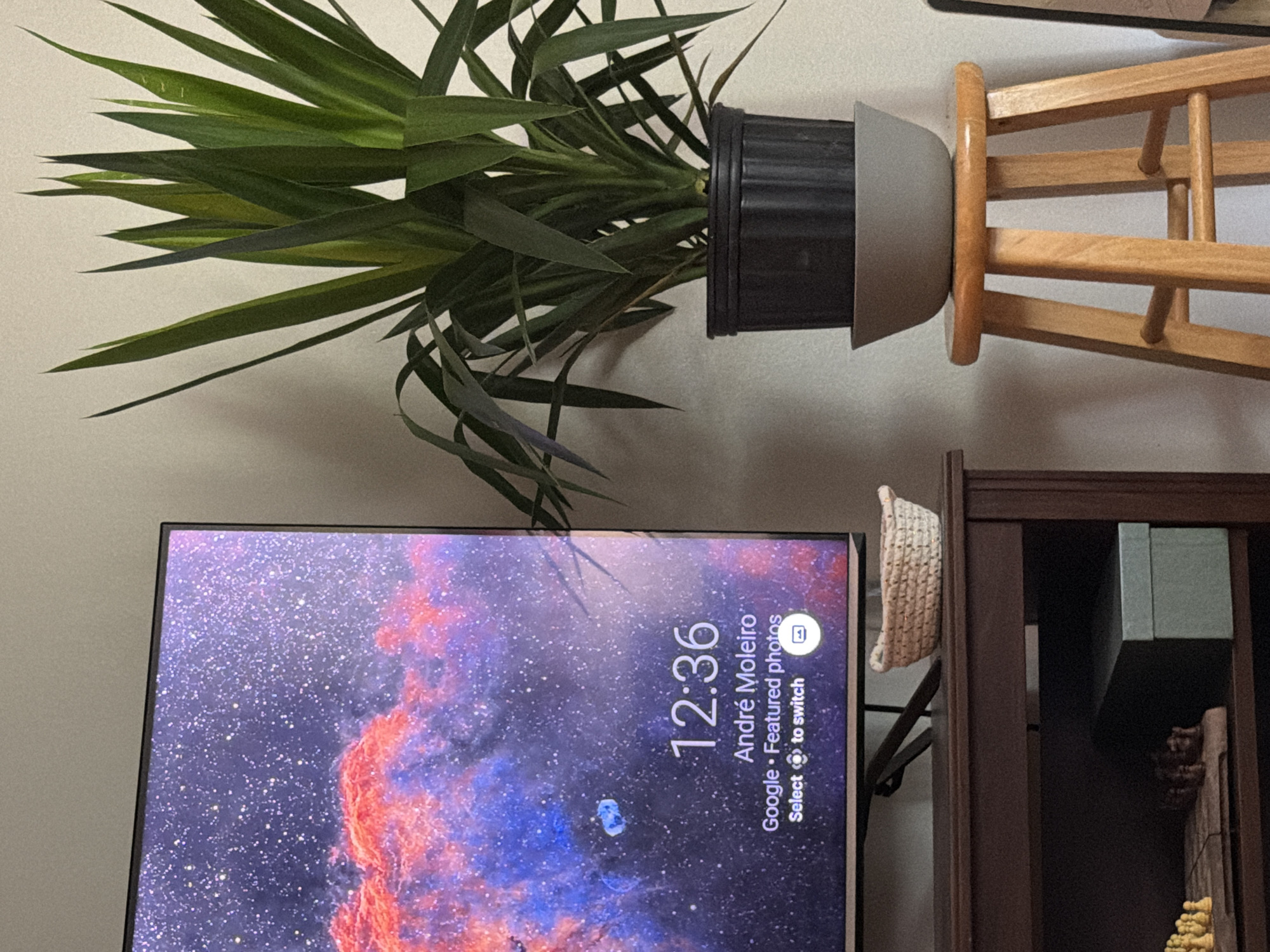 The image shows a section of an interior scene. A green plant in a gray and black pot is resting on a light wooden stand, which is mounted on a light-colored wall. Below the plant is a TV screen displaying a digital clock that reads 12:36. The screen's background is an image of a nebula. To the right of the TV screen is a dark wooden cabinet or shelf, partially visible, with a woven basket hanging from it. There is no specific indication of Morton Grove, United States of America, visible in the scene. The image focuses on domestic objects and displays no people or actions.