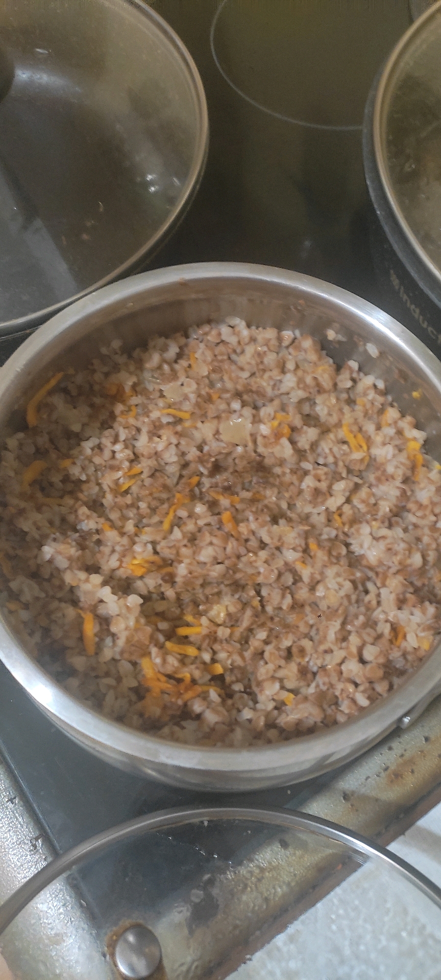 The image shows a close-up view of a pot of cooked buckwheat with shredded carrots on a stovetop. The buckwheat appears to be a staple food in Russia. The scene is likely in a domestic setting in the city of Bolshoy Kamen, Russia. The stovetop is dark and appears to be an induction hob. There are other pots or pans with lids visible in the background, suggesting a meal is being prepared or has recently been cooked. The lighting is artificial and somewhat dim.