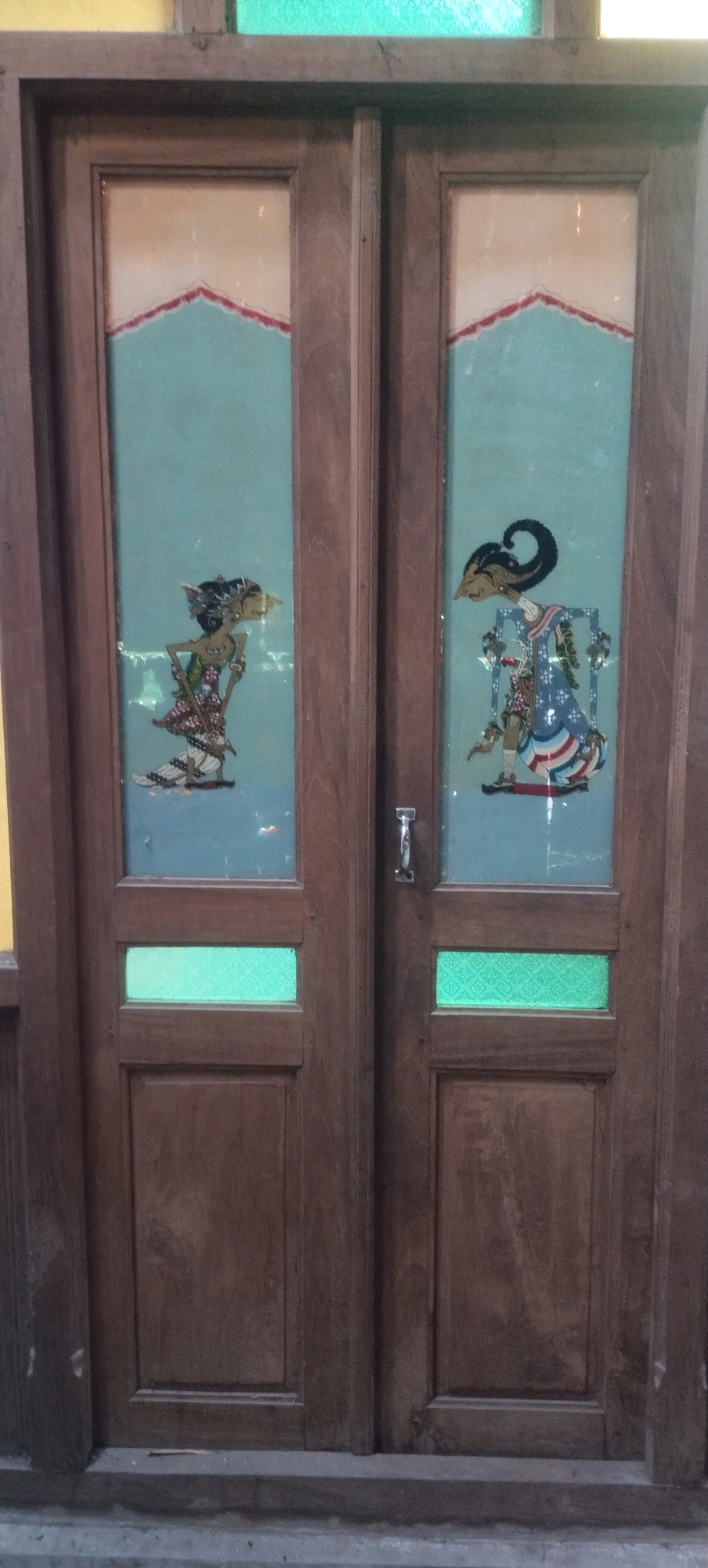 The image shows a closed wooden double door, likely an entrance. The doors are made of dark wood and are divided into several panels. The upper panels of each door feature a decorative element: a stylized silhouette of a figure against a light blue background, bordered by a triangular shape with red and white detailing at the top. The figure on the left appears to be female with elaborate hair and clothing. The figure on the right is also stylized, with a prominent headdress and patterned attire. Below these decorative panels, each door has two rectangular panels, the upper one of which is a translucent green material, possibly stained glass or patterned plastic, and the lower panel is solid wood. A silver-colored handle is visible on the right door. The doors are set within a wooden frame, and a hint of green and yellow colored glass is visible at the very top of the frame. The setting appears to be outdoors, with a concrete base visible at the bottom of the doors. The overall impression is of a traditional or artisanal aesthetic. The location is specified as Daleman, Indonesia, suggesting the cultural context of the artwork on the doors.