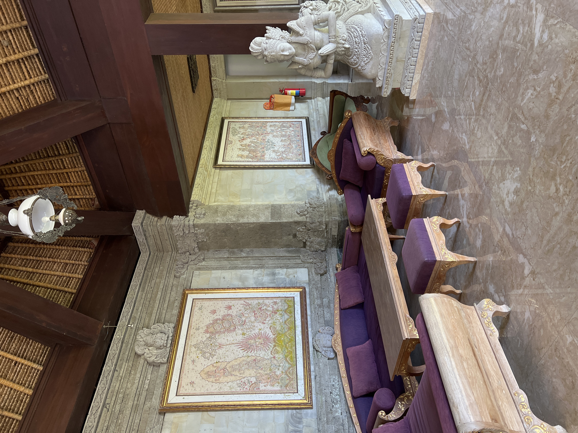 The image depicts an interior scene in Kedewatan, Indonesia, featuring ornately decorated furniture, artwork, and architectural details. The furniture set includes a long, upholstered sofa with purple cushions, accompanied by matching chairs and tables with ornate, gold-colored trim. Two framed paintings hang on the wall, depicting intricate, detailed scenes. A large white statue stands on a platform in the background. The room's architectural features include a thatched roof, dark wooden beams, and carved stone walls. A chandelier hangs from the ceiling. The floor is covered in marble tiles. There are no people visible in the scene.