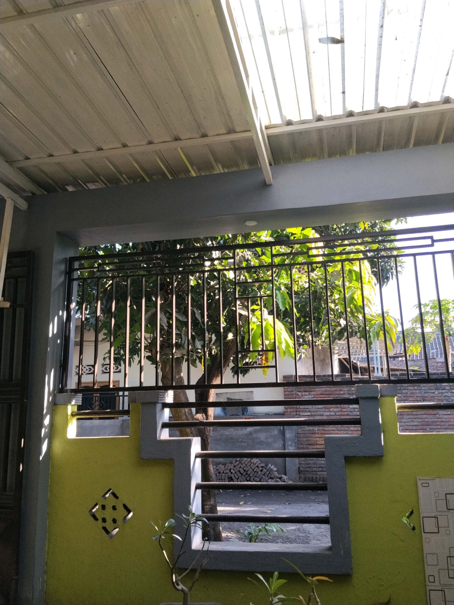 The image depicts a view through a metal fence and an overhang in what appears to be a residential outdoor space in Korleko Selatan, Indonesia. The roof above is constructed of corrugated metal, with some sections appearing translucent, allowing sunlight to filter through, casting striped shadows on a grey support beam and the yellow wall below. The fence itself is black metal, with a decorative geometric pattern on the upper section. Behind the fence, a tree with lush green leaves is visible, its trunk and branches illuminated by the sun. Beyond the tree, a building with red brick accents and white walls can be seen, along with what looks like a stack of bricks. The yellow wall in the foreground features a diamond-shaped cutout with smaller square openings, and a small plant is growing near the bottom of the frame. The overall scene suggests a bright, sunny day.