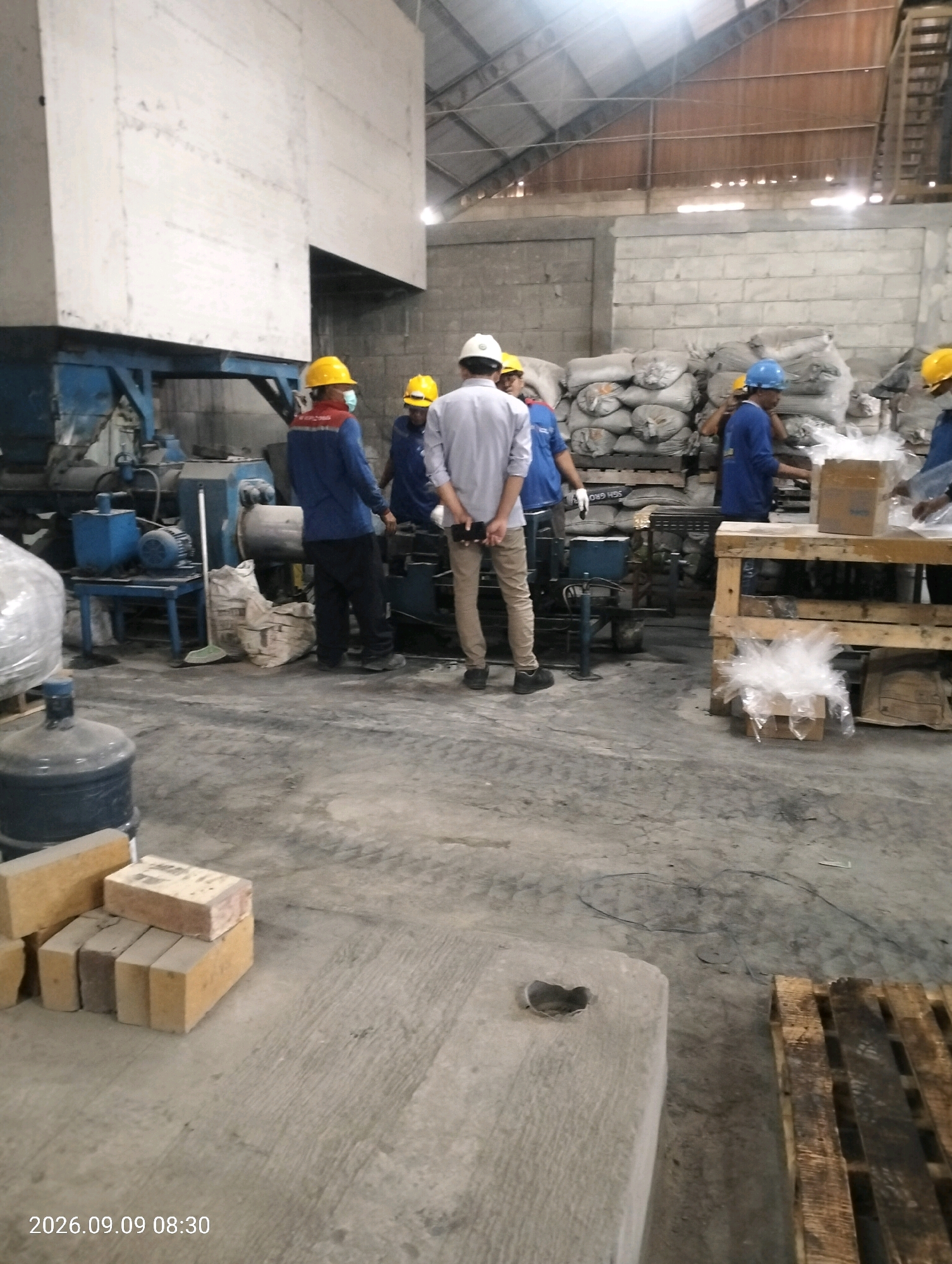 A group of workers, wearing hard hats, are in an industrial warehouse. The warehouse has a concrete floor and brick walls. There are stacks of bags and pallets of goods in the background. In the foreground, there is a large blue water cooler and a stack of bricks. One worker is wearing a blue shirt and black pants, another worker is wearing a white shirt and khaki pants. The scene appears to be in Bambe, Indonesia, as indicated by the date stamp at the bottom of the image.