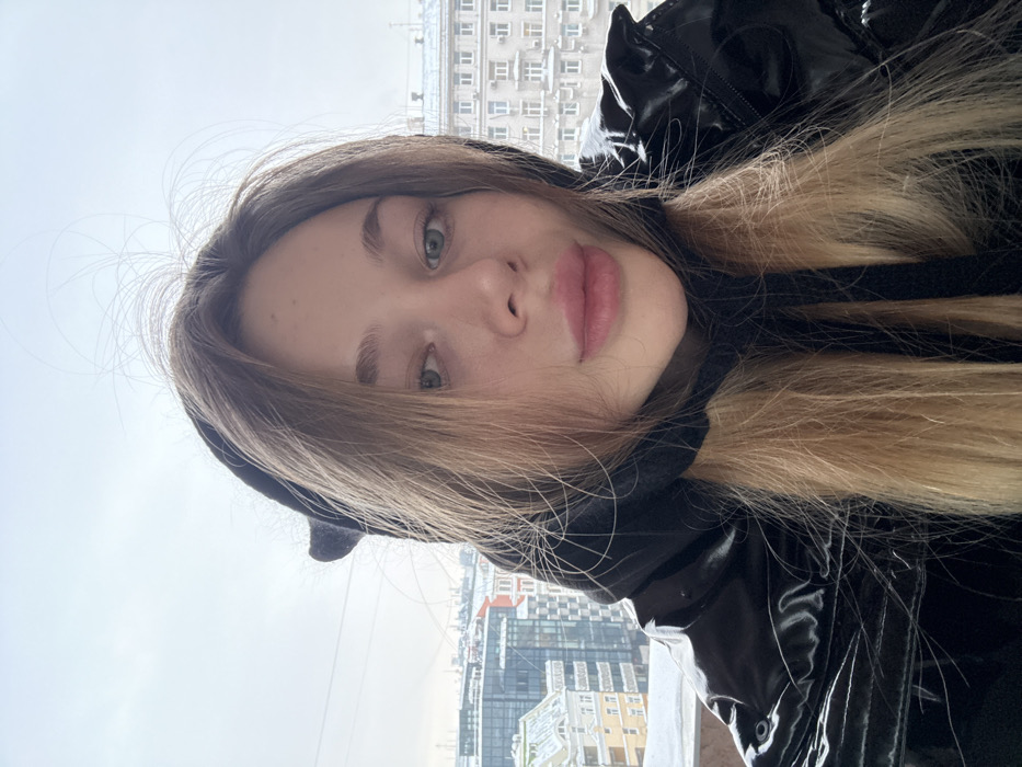 A young woman takes a selfie indoors with a city backdrop. She wears a black jacket and a hood with small ears. Her long blonde hair is partially covering her face. She faces the camera at an angle with a slight smirk. The sky outside appears overcast and the city buildings in the background are typical high rise structures.