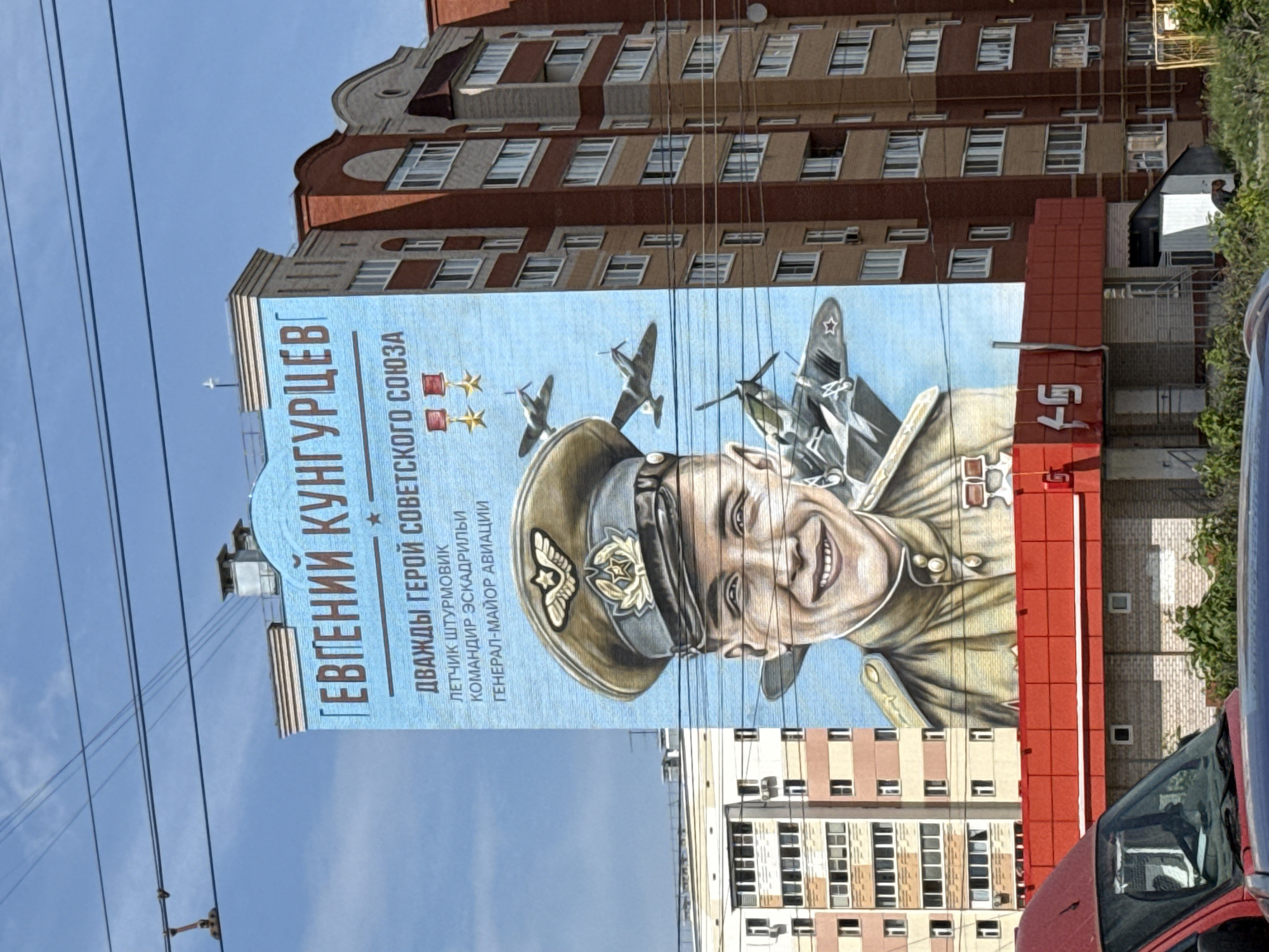 The image shows a mural on the side of a building in Izhevsk, Russia. The mural depicts Evgeniy Kungurtsev, a Soviet pilot and twice Hero of the Soviet Union. Text on the mural describes Kungurtsev as a fighter pilot, squadron commander, and General-Major of Aviation. The mural also features images of airplanes. The building is brick with multiple floors and windows, with a red decorative element on the side of the building. There are other apartment buildings in the background. Power lines are visible in the foreground of the image. A small section of a red car is also in the foreground.